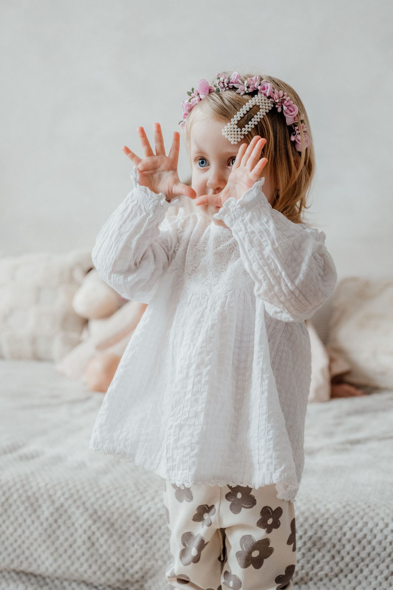 Cute little girl in white lace dress and floral hair clips raising hands playfully on a bed.