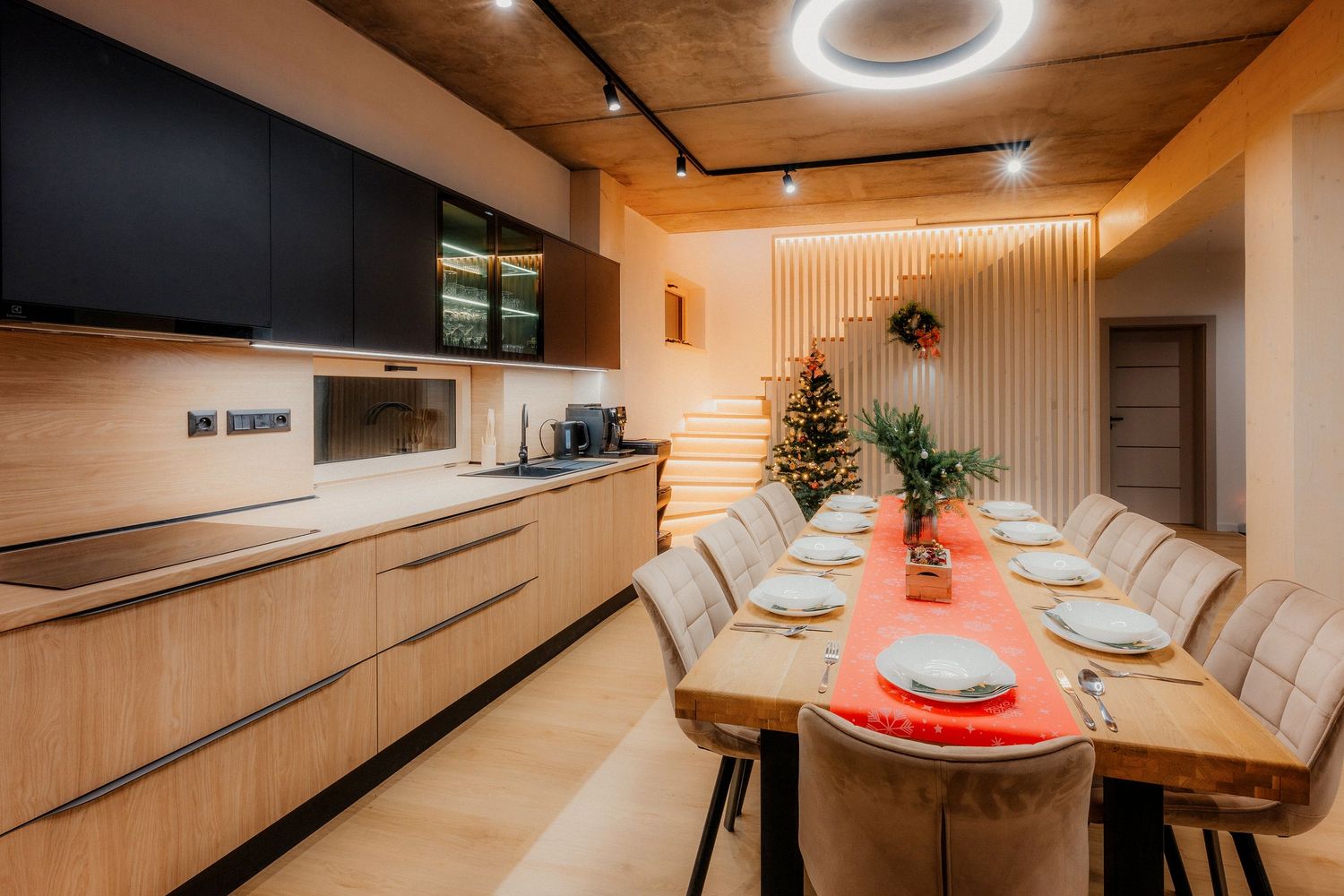 Modern open-plan kitchen with wood cabinets, long dining table set for Christmas dinner with festive decor.