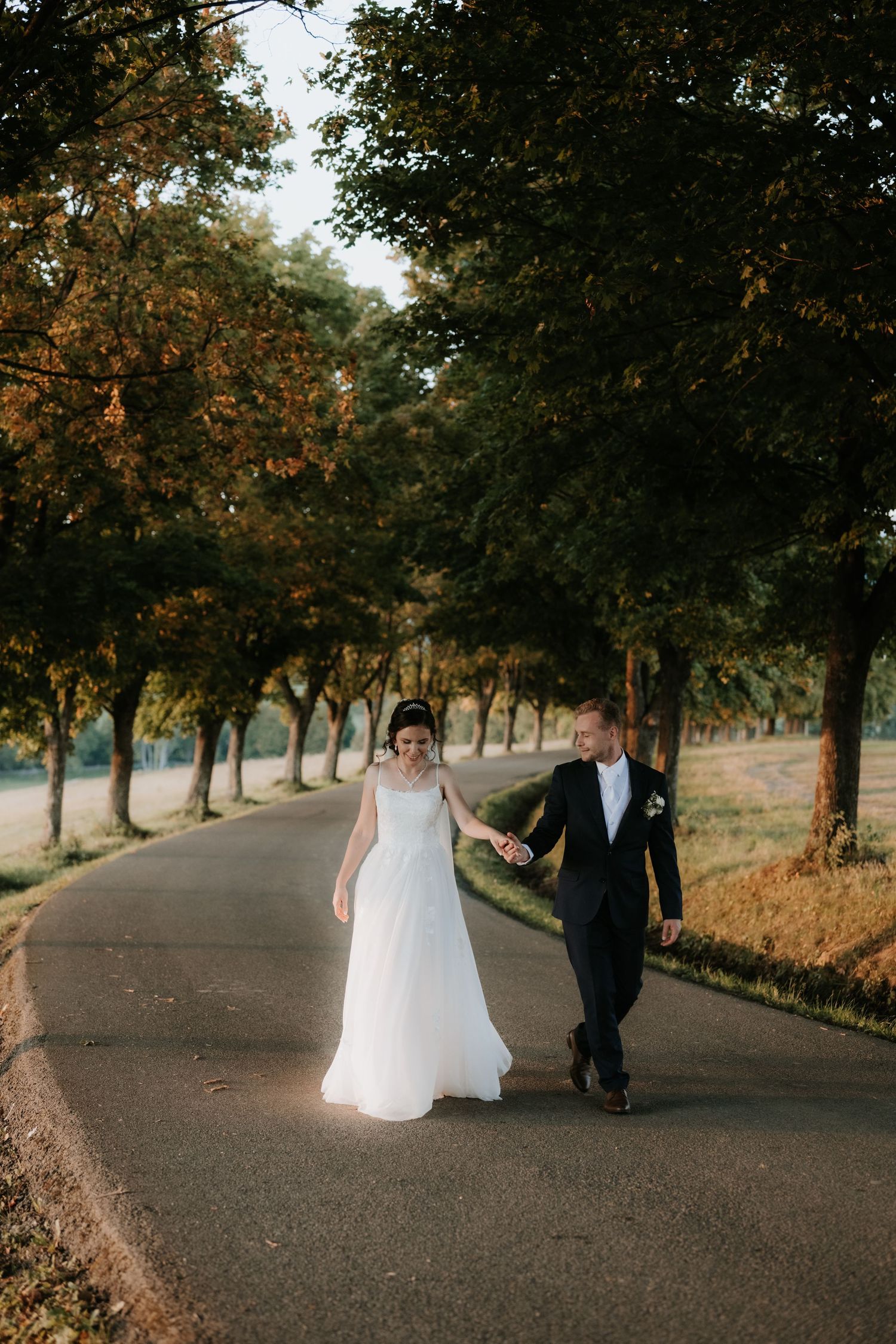 A bride and groom walk hand in hand down a tree-lined road in golden hour light on their wedding day.