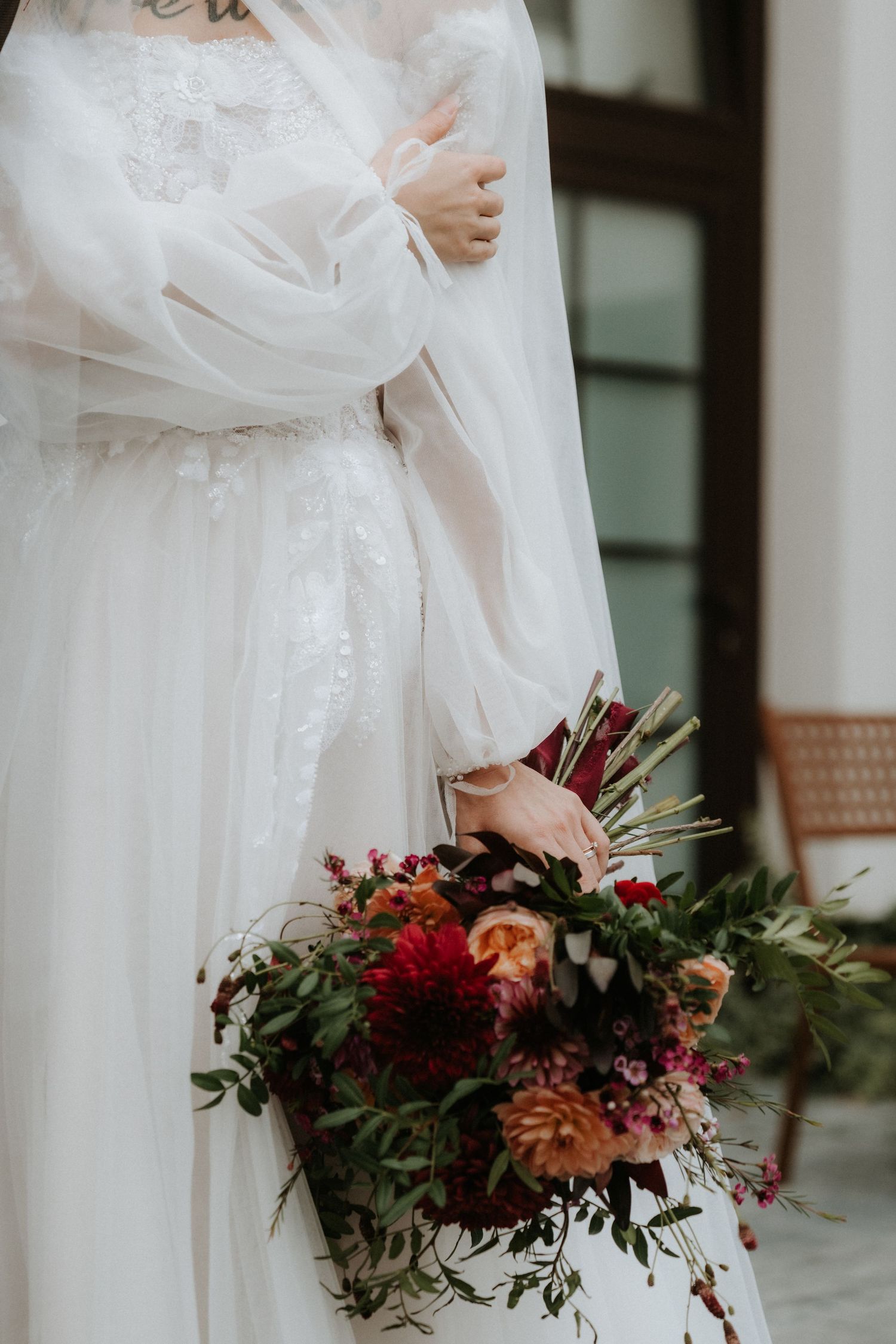 Bride in white lace gown holding a lush bridal bouquet with deep red, pink, and orange flowers with greenery.