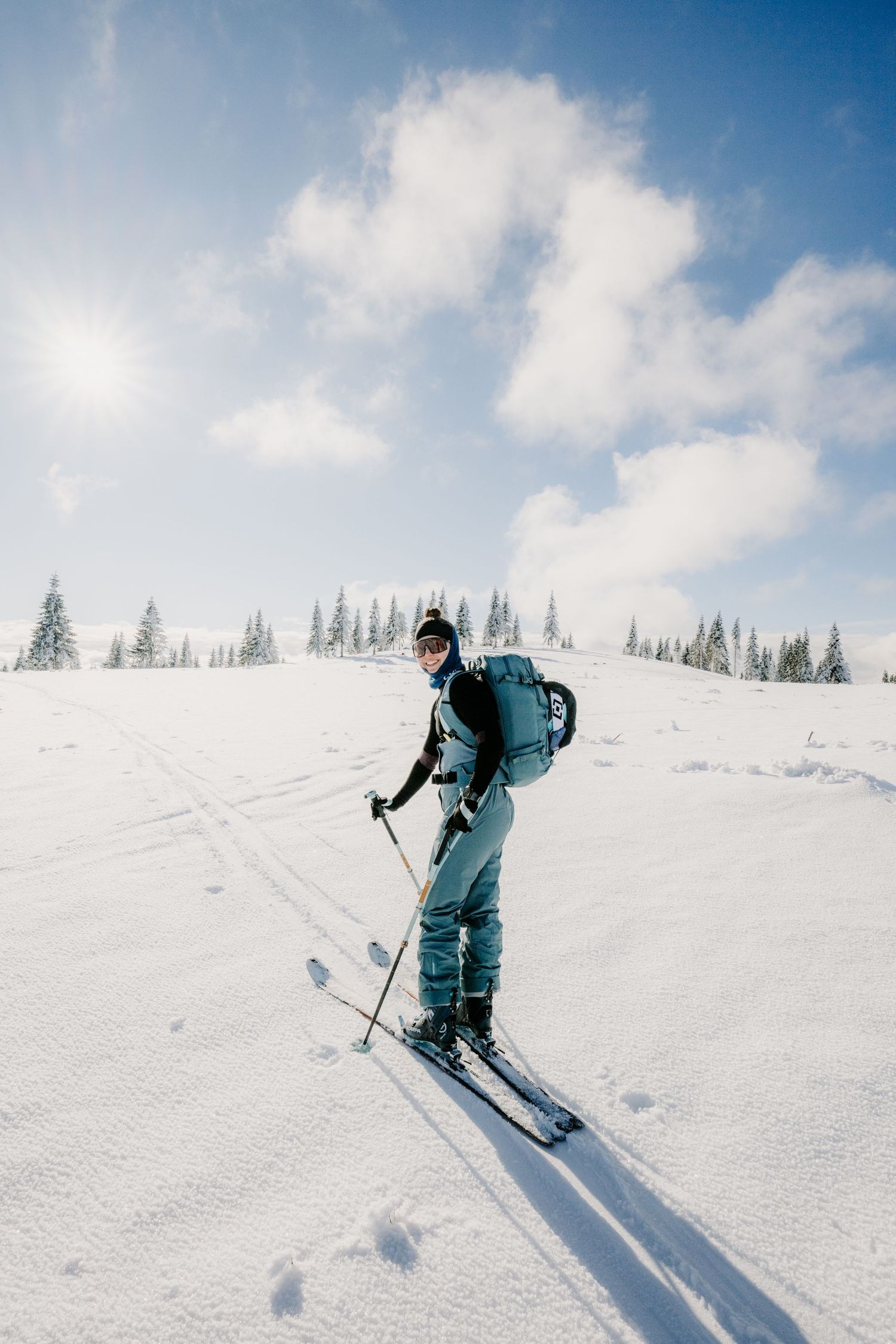 A smiling skier with a backpack poses on a sunny snow-covered slope with pine trees in the distance.