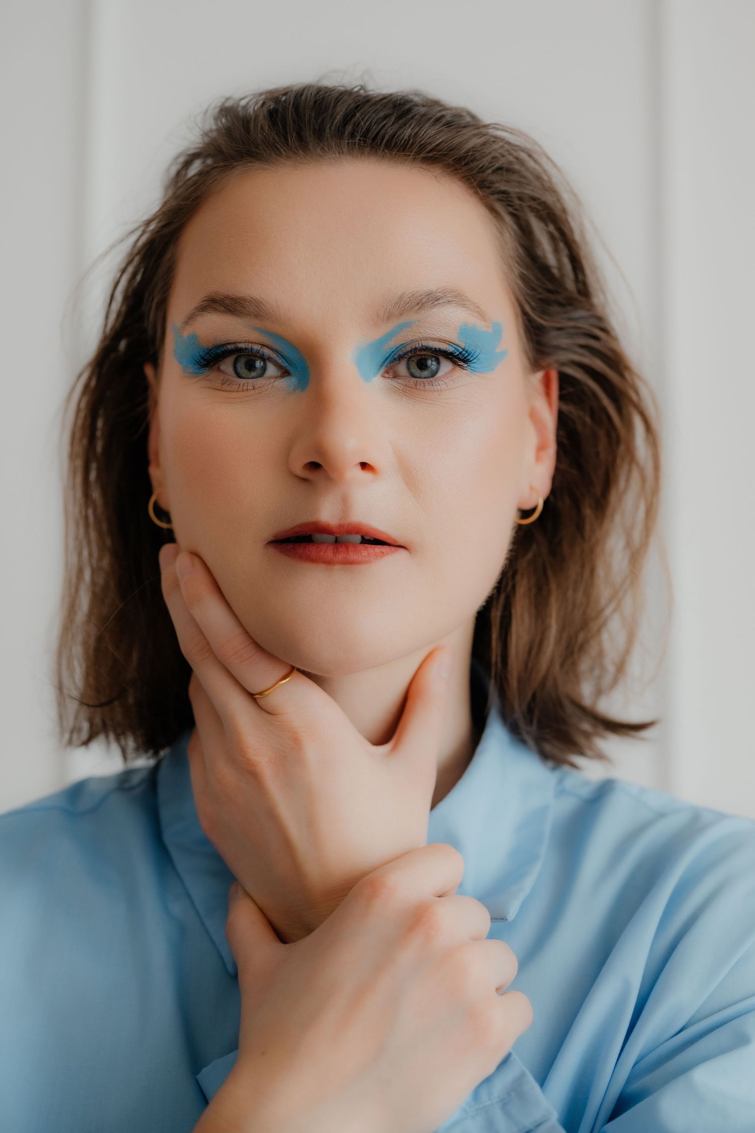 Woman with bold blue eye makeup and red lips in blue shirt touching her chin.