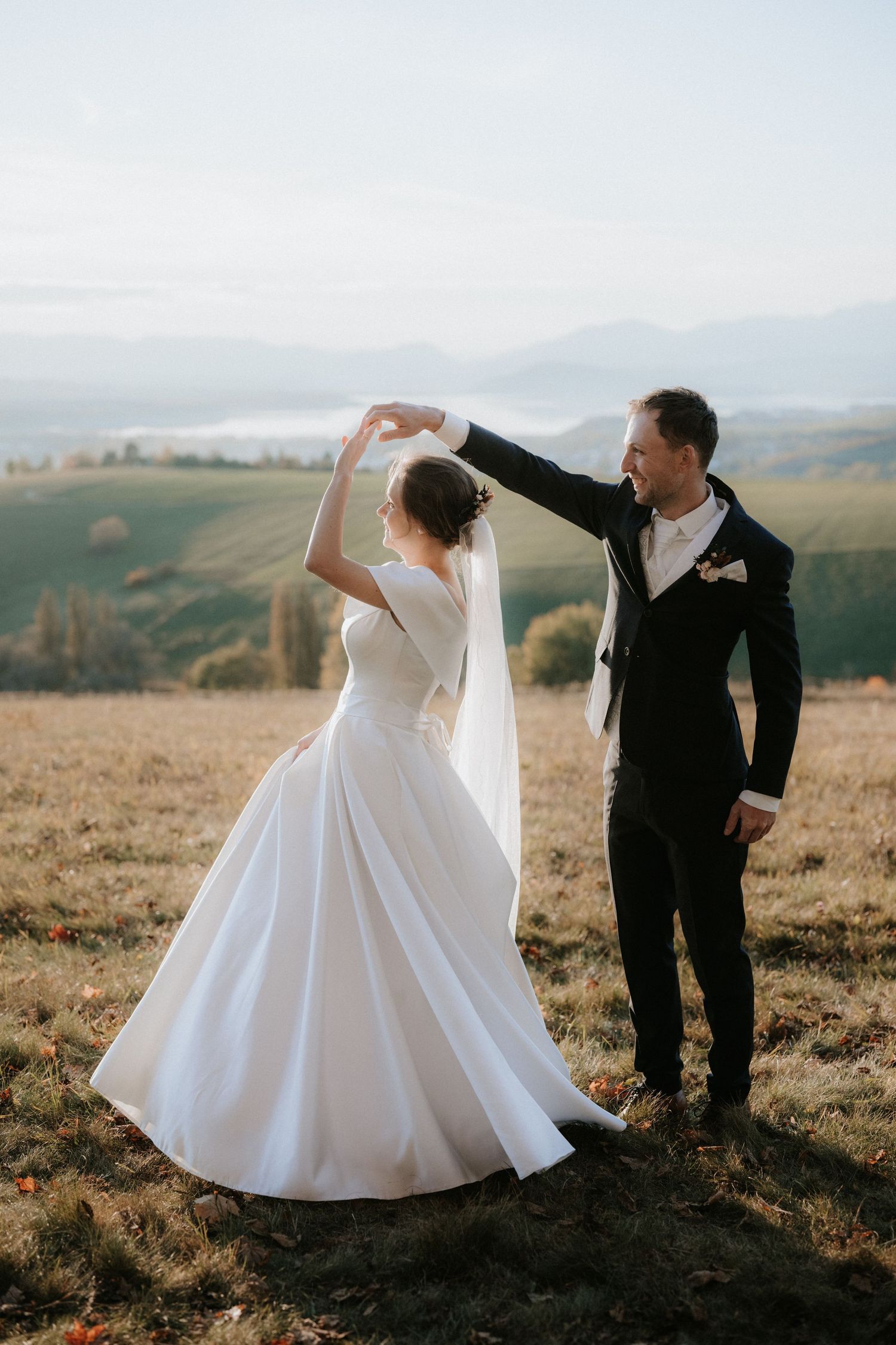 Bride and groom dancing in field at golden hour with mountain landscape in background.