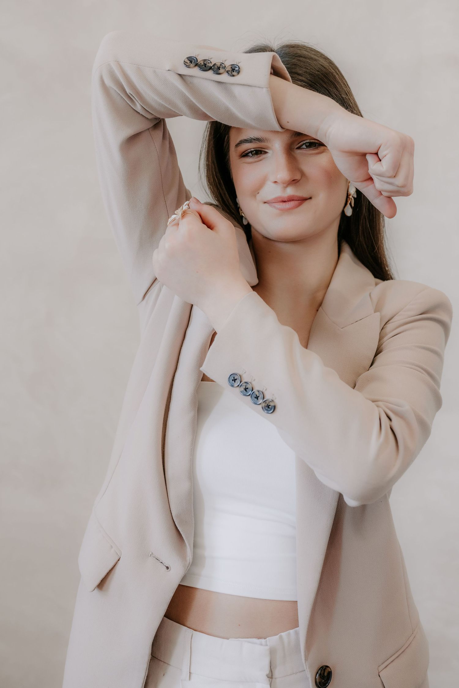 Brunette woman in beige blazer and white crop top posing playfully against neutral background.