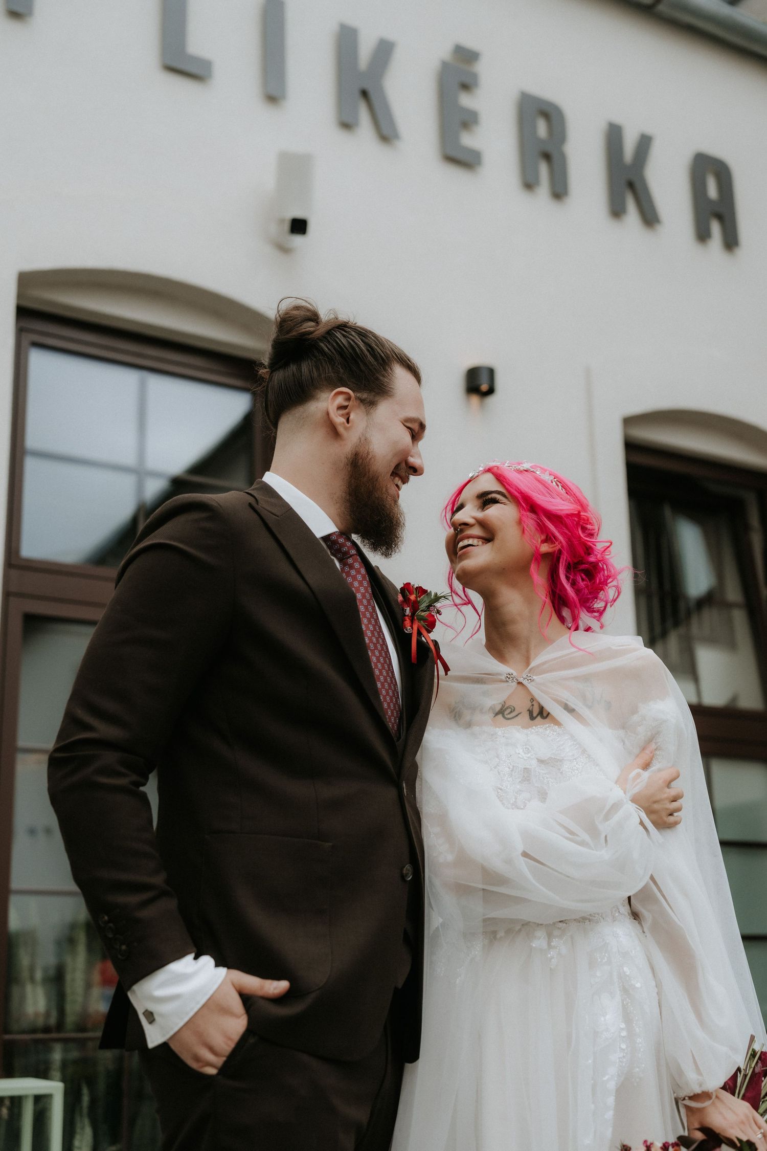 Smiling pink-haired bride in white dress and tulle cape gazing at bearded groom outside venue.