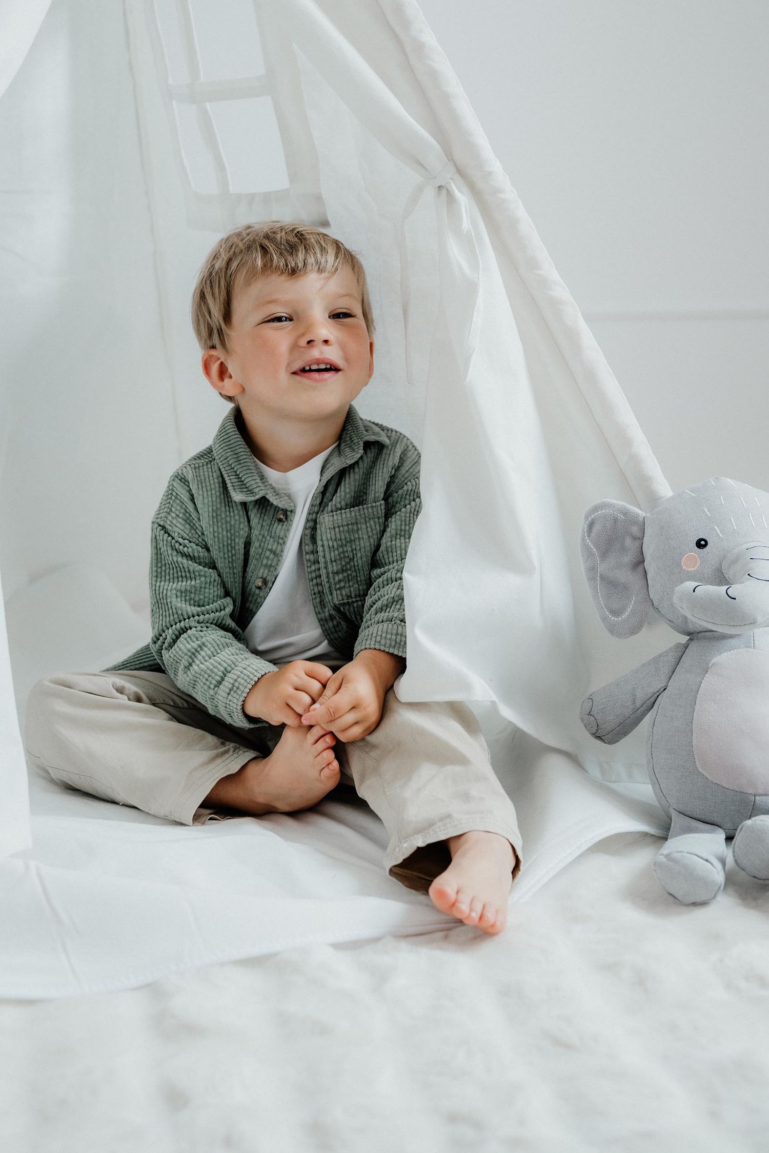 Happy toddler boy sitting barefoot near white teepee tent with elephant plush toy.