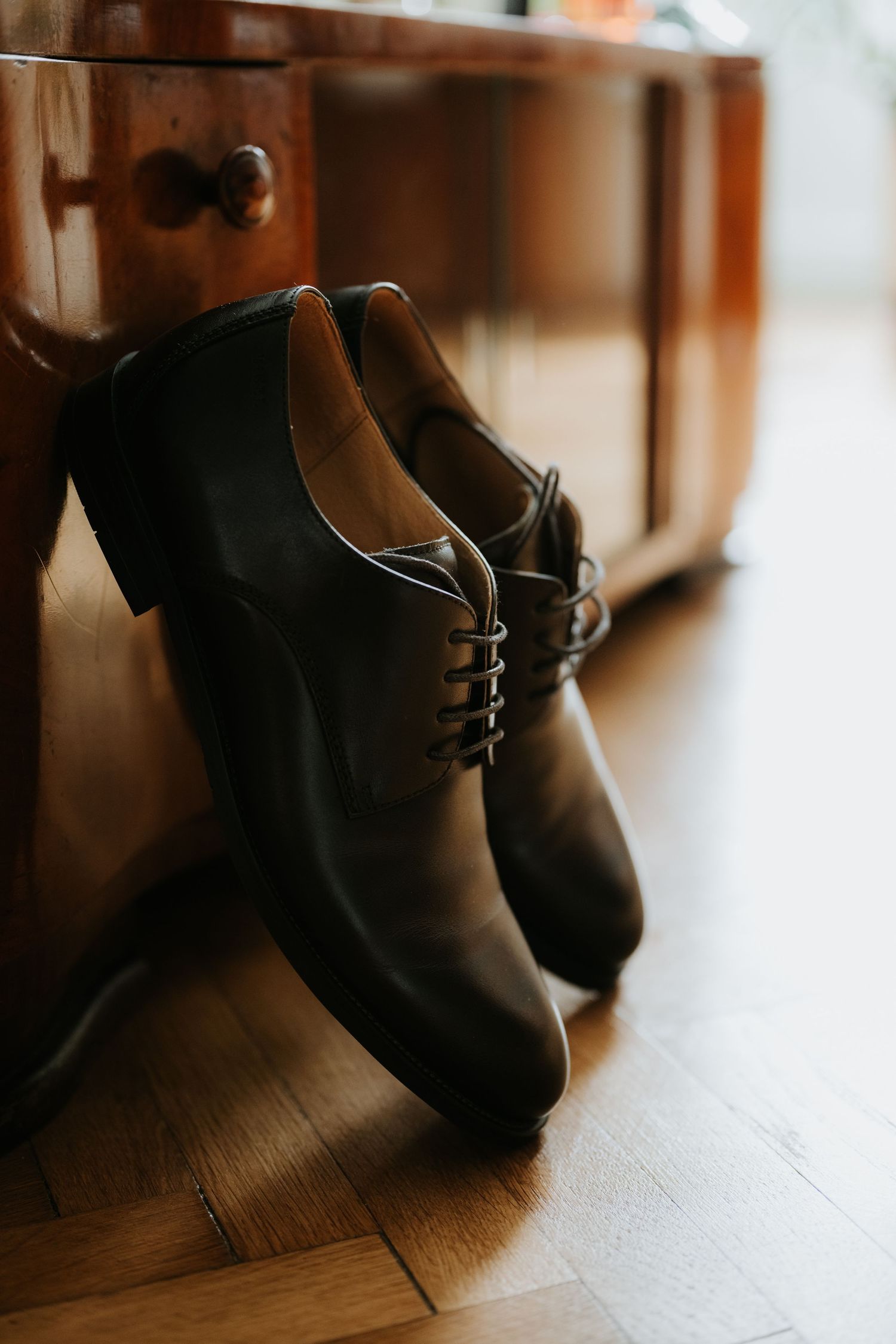 Elegant black leather Oxford dress shoes leaning against a wooden dresser on a hardwood floor.