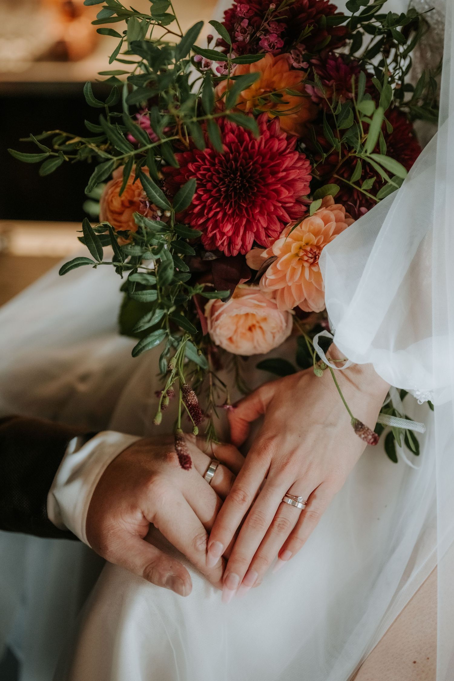 Bride and groom holding hands with wedding rings visible, beside a vibrant bouquet of red dahlias and orange roses.