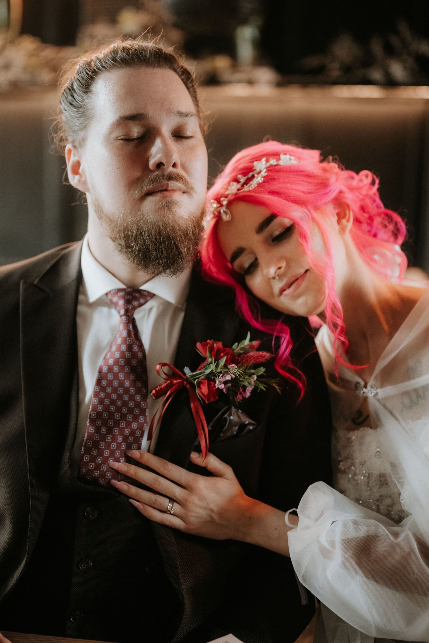 Bride with pink hair leans on groom in suit at wedding, holding red floral bouquet.