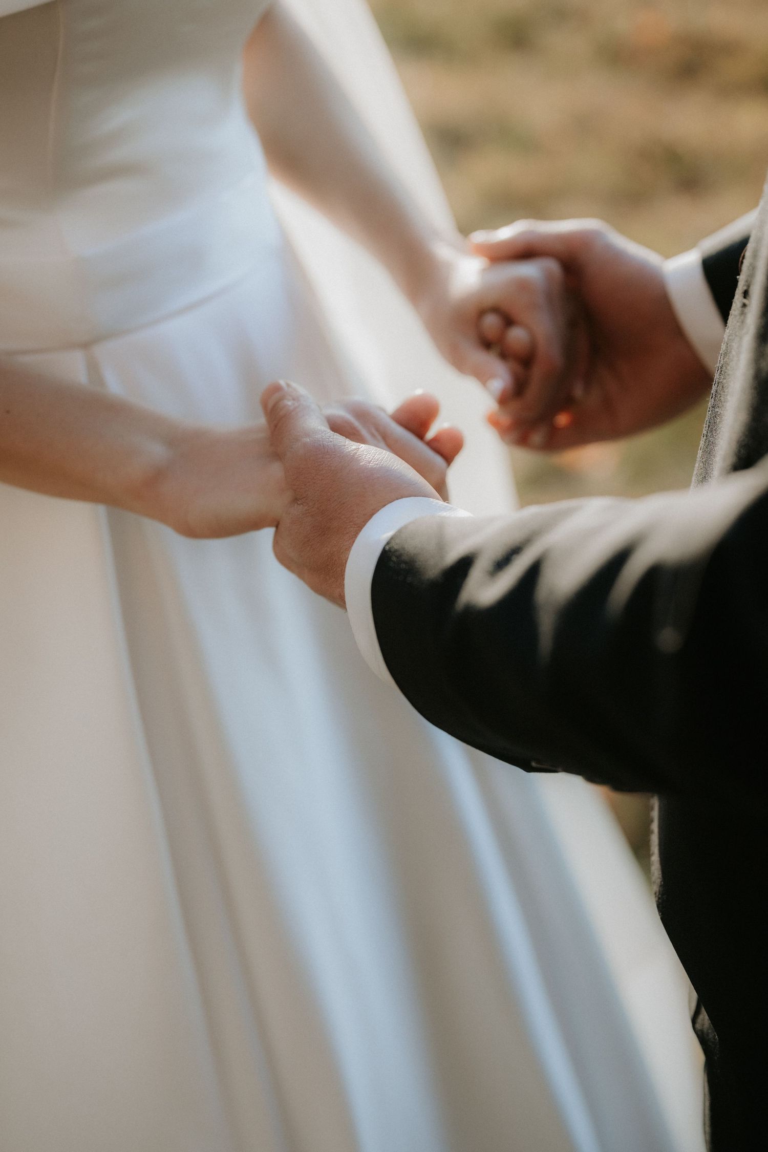 Bride and groom holding hands during wedding ceremony, close-up of white bridal gown and dark suit.