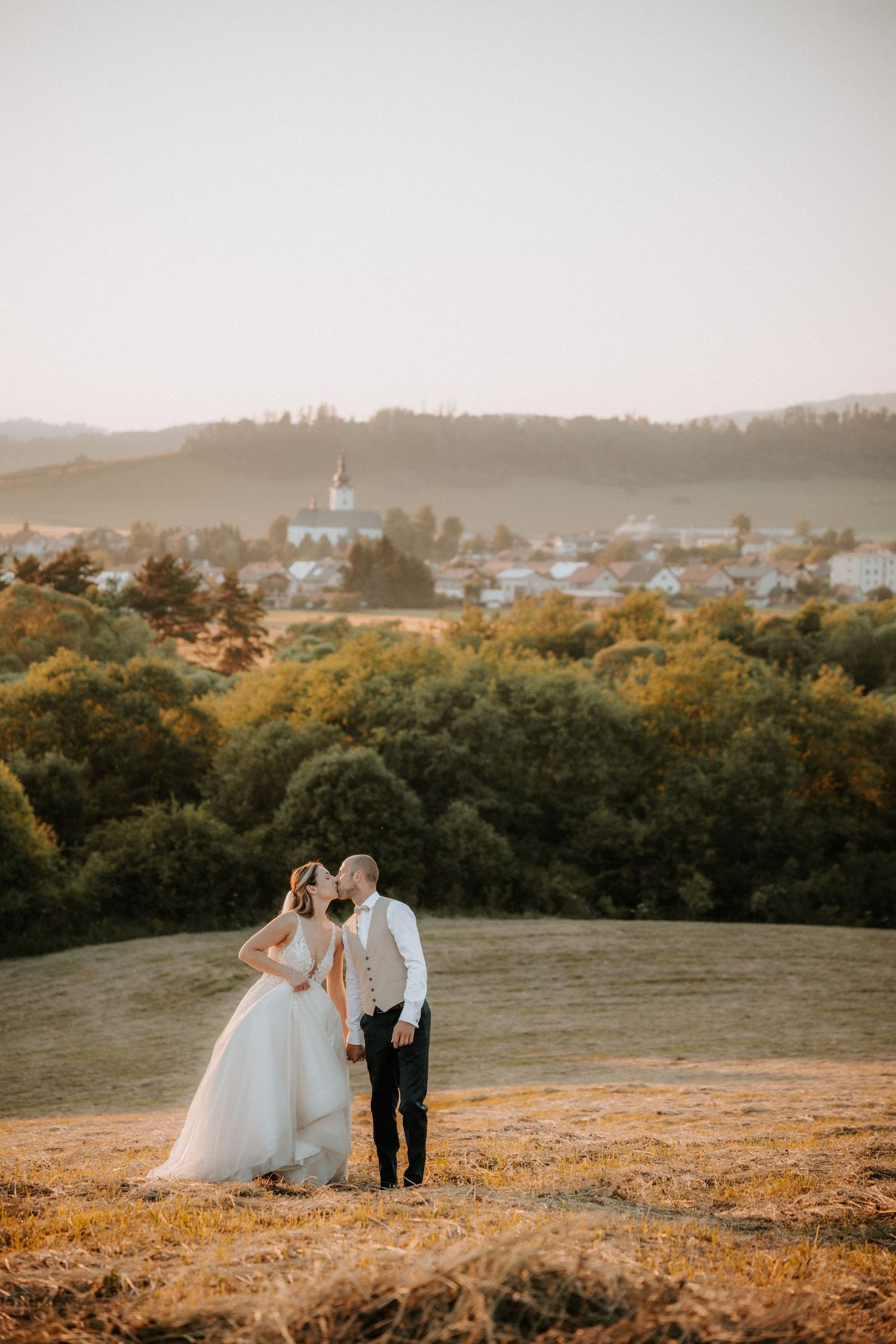 Wedding couple kissing on a sunlit hillside with a charming village, lush green trees, and mountains in background.