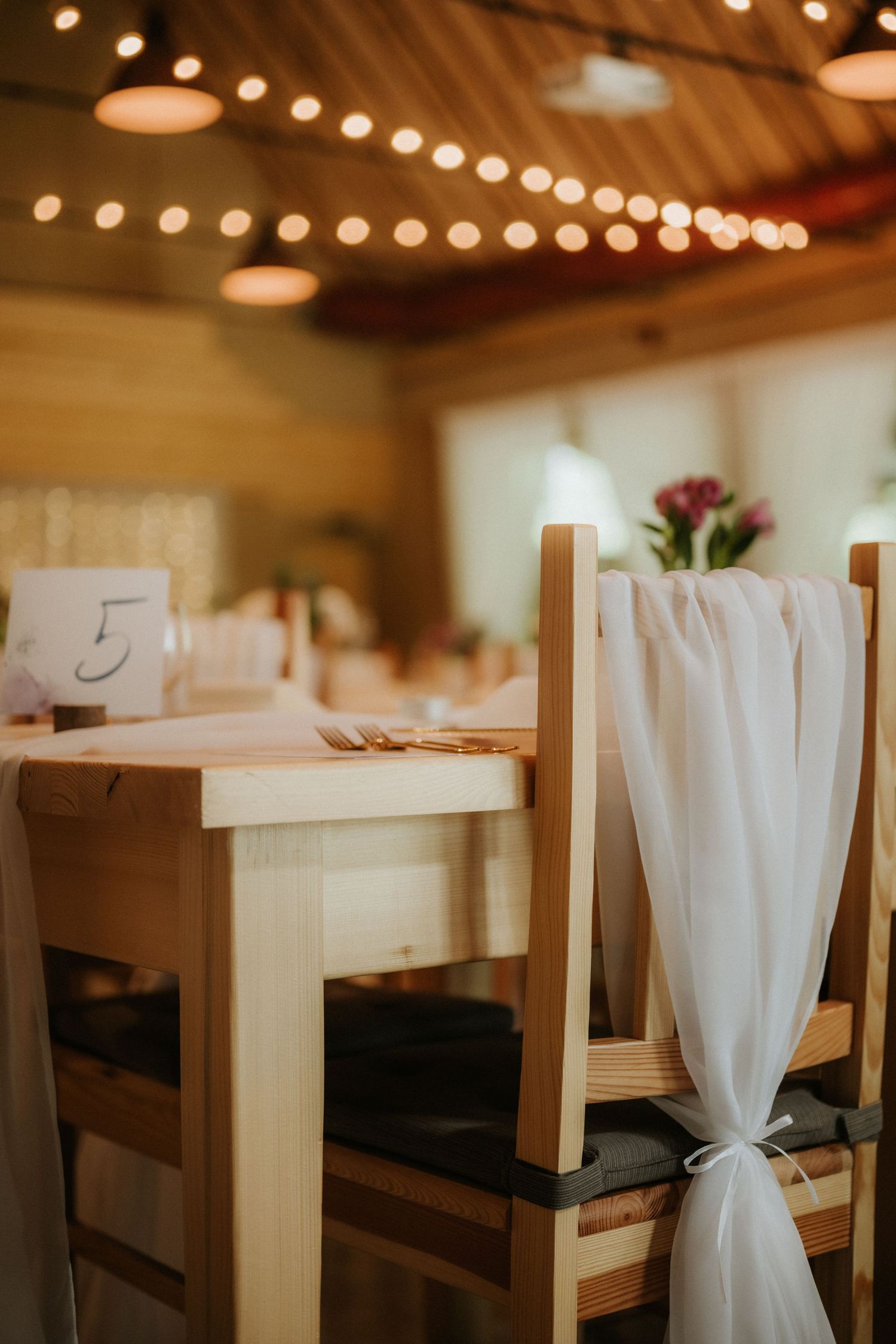 Rustic wedding reception venue with wooden chairs draped in white tulle, string lights overhead, and table number five visible.