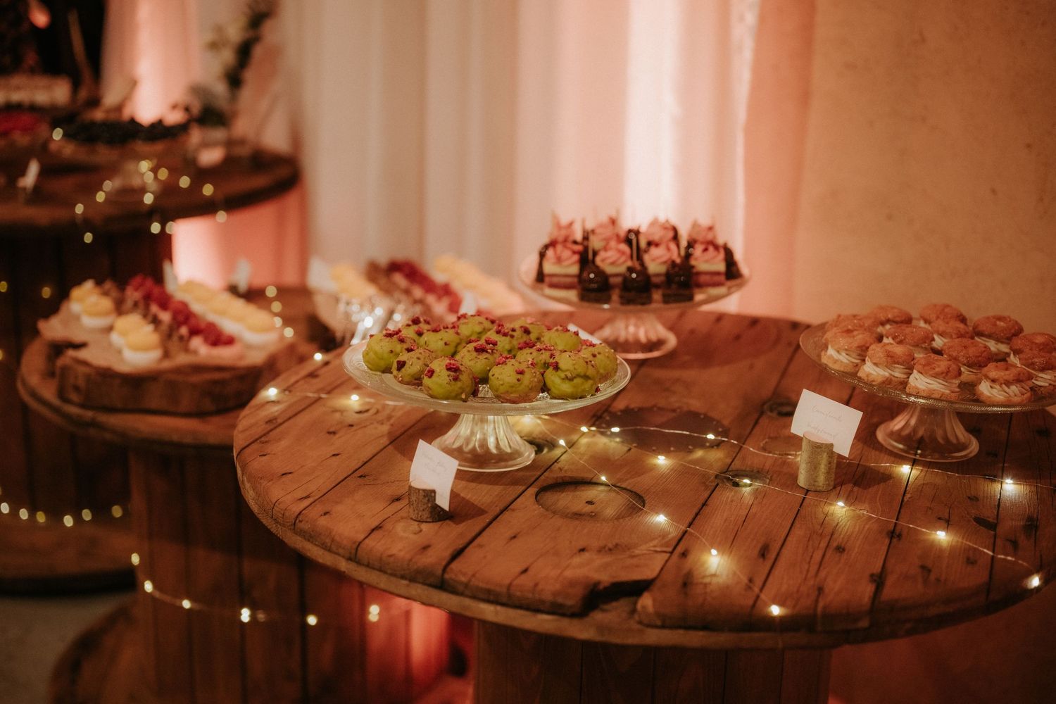 Rustic wooden spool tables adorned with string lights display an elegant dessert spread at a warmly lit wedding reception.