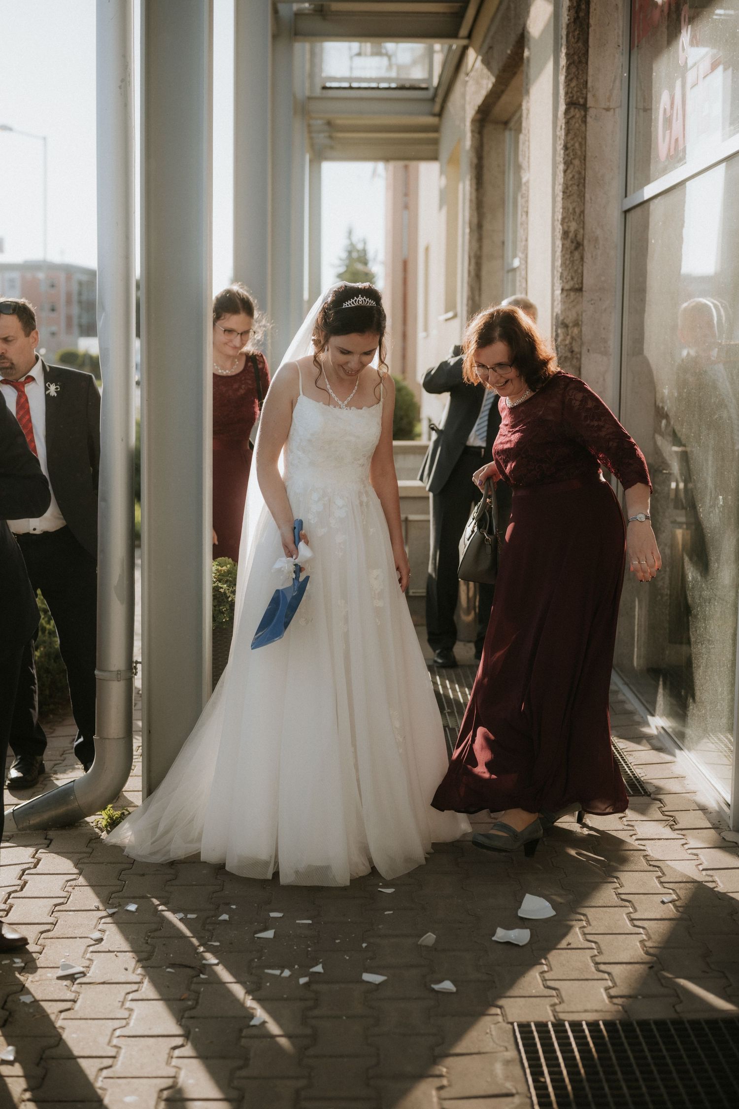 Bride in white gown walks with guests in burgundy dresses on a sunny sidewalk during a wedding celebration.