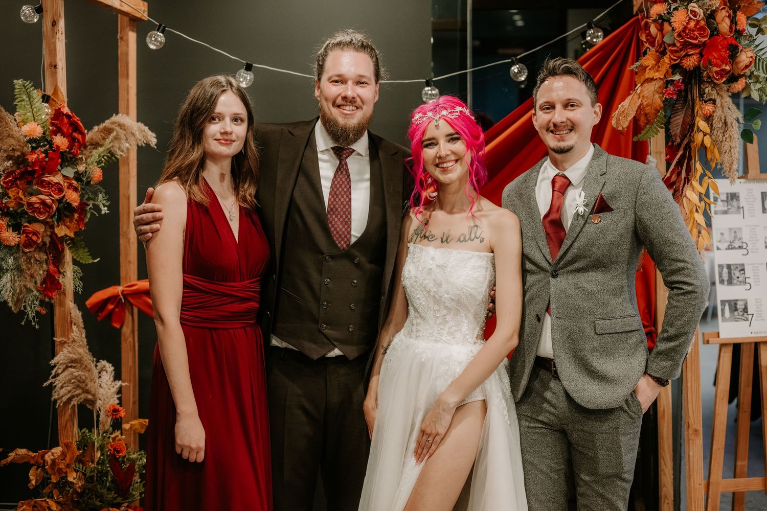 Wedding party of four posing together, bride in white gown with pink hair, bridesmaid in red dress, two groomsmen.
