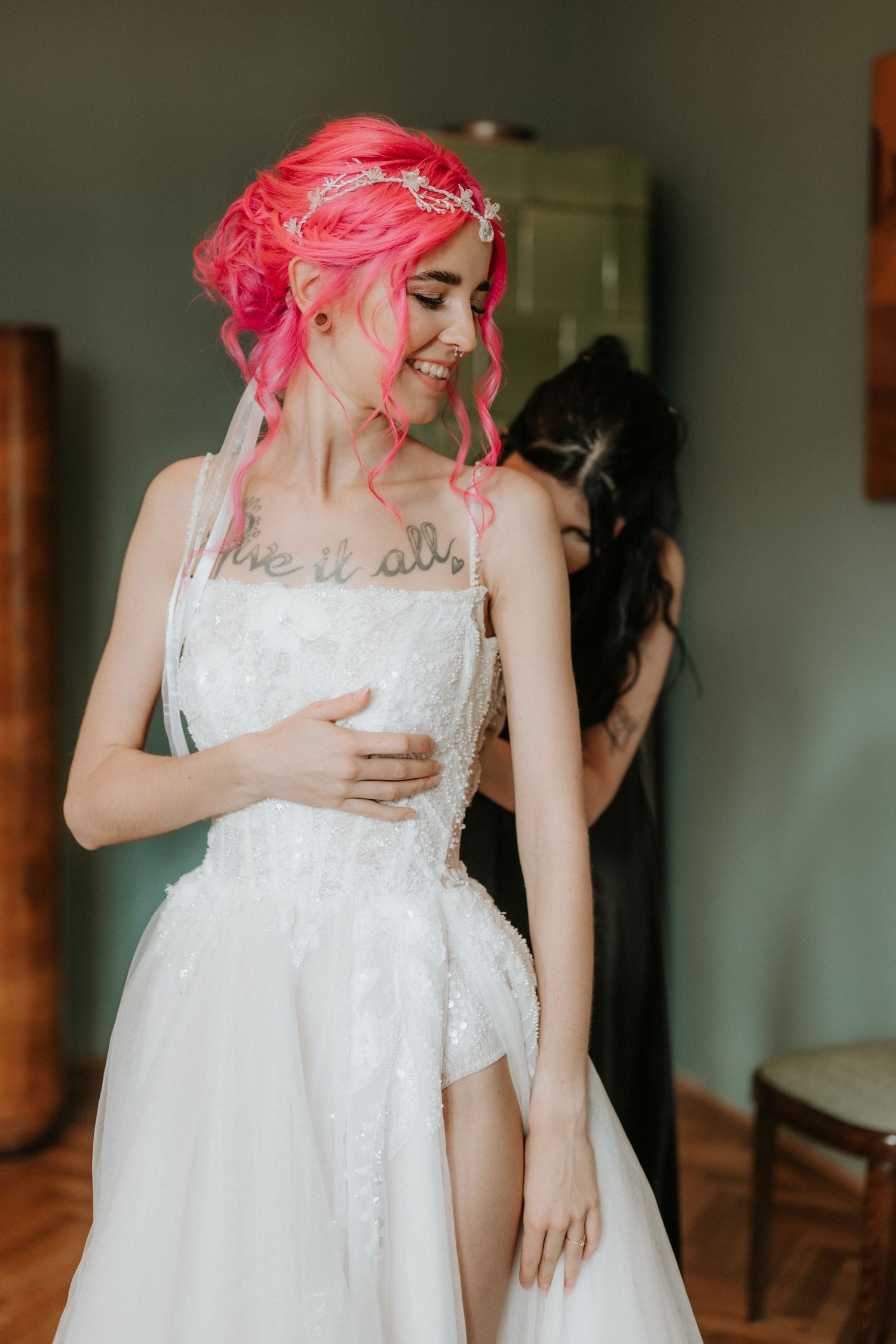 Pink-haired bride in white lace wedding dress smiles as bridesmaid helps fasten her gown indoors.