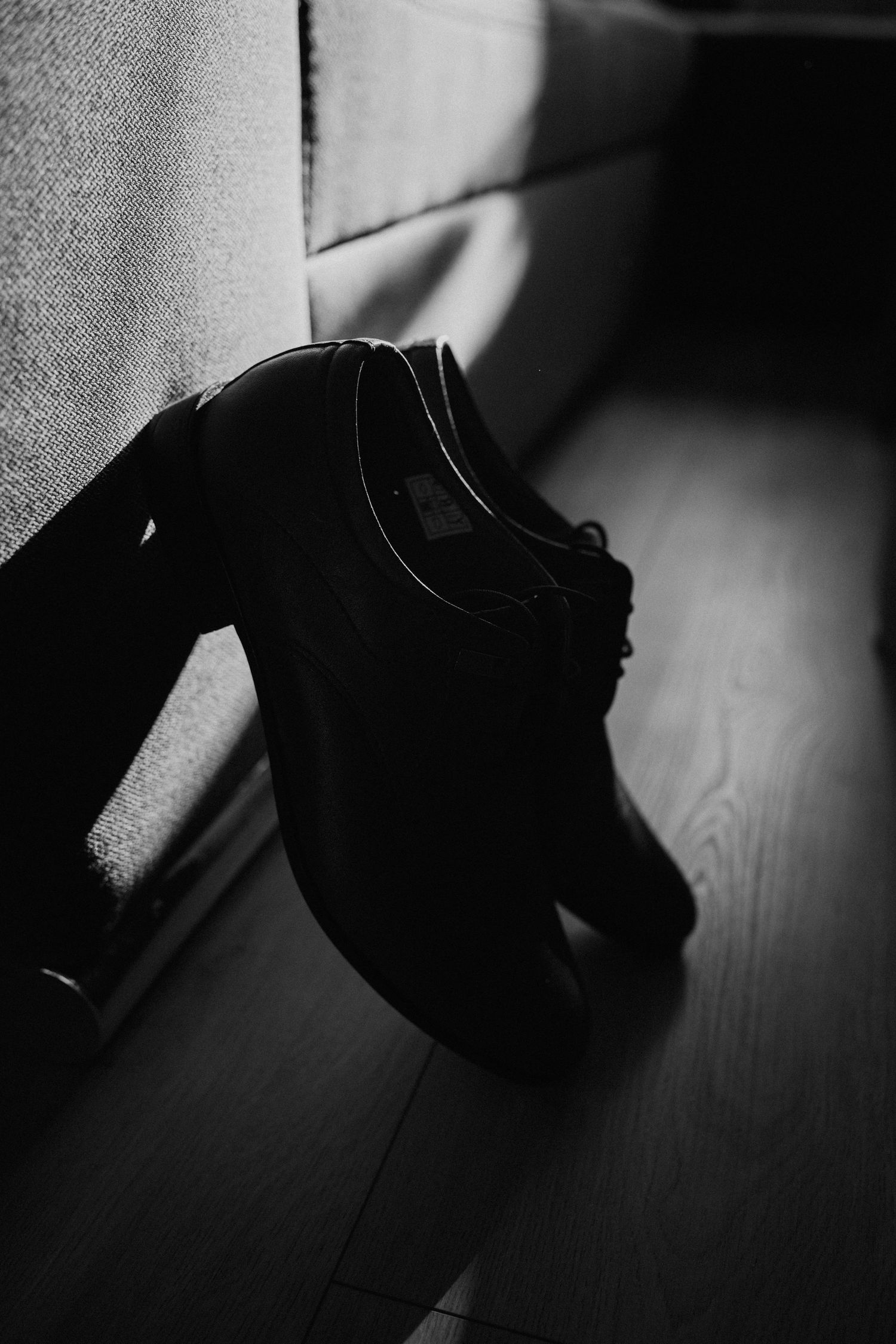 Black and white photo of elegant dress shoes resting against a sofa on a wooden floor.