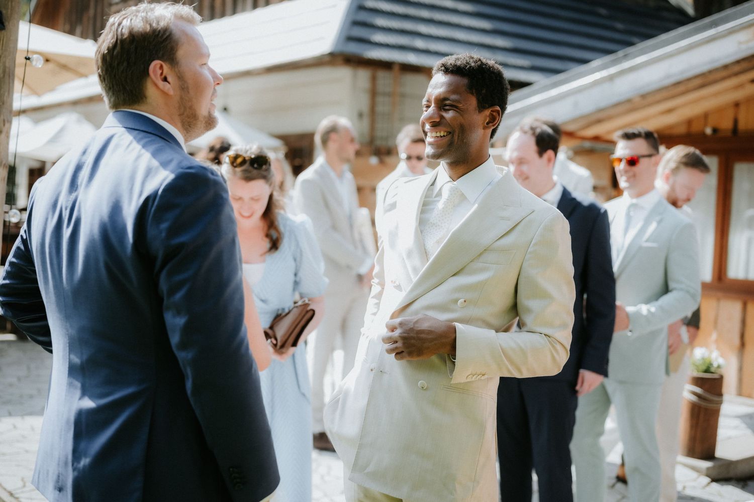 Two men in elegant suits conversing at an outdoor wedding reception with guests in background.