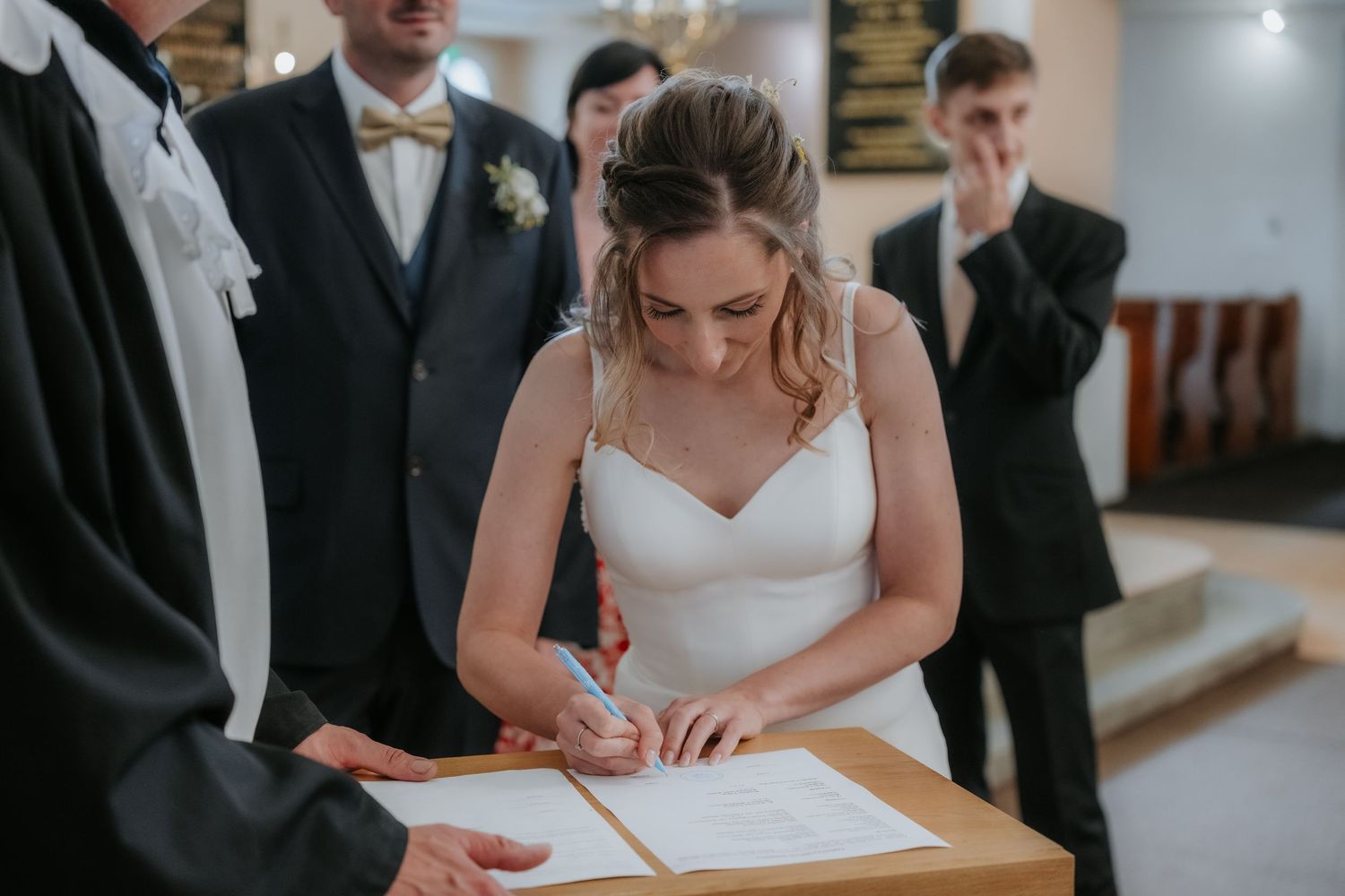 Bride in white gown signing wedding register at ceremony table with groom and guests in background.