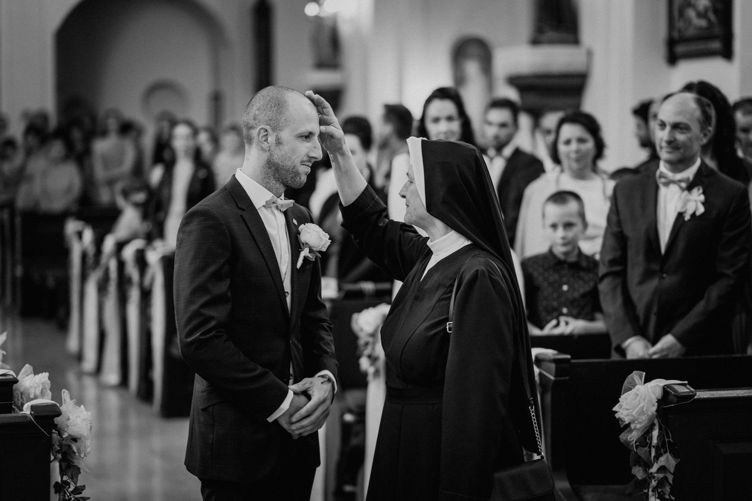 A nun blesses a groom in a black suit during a church wedding ceremony, with guests seated in pews behind them.