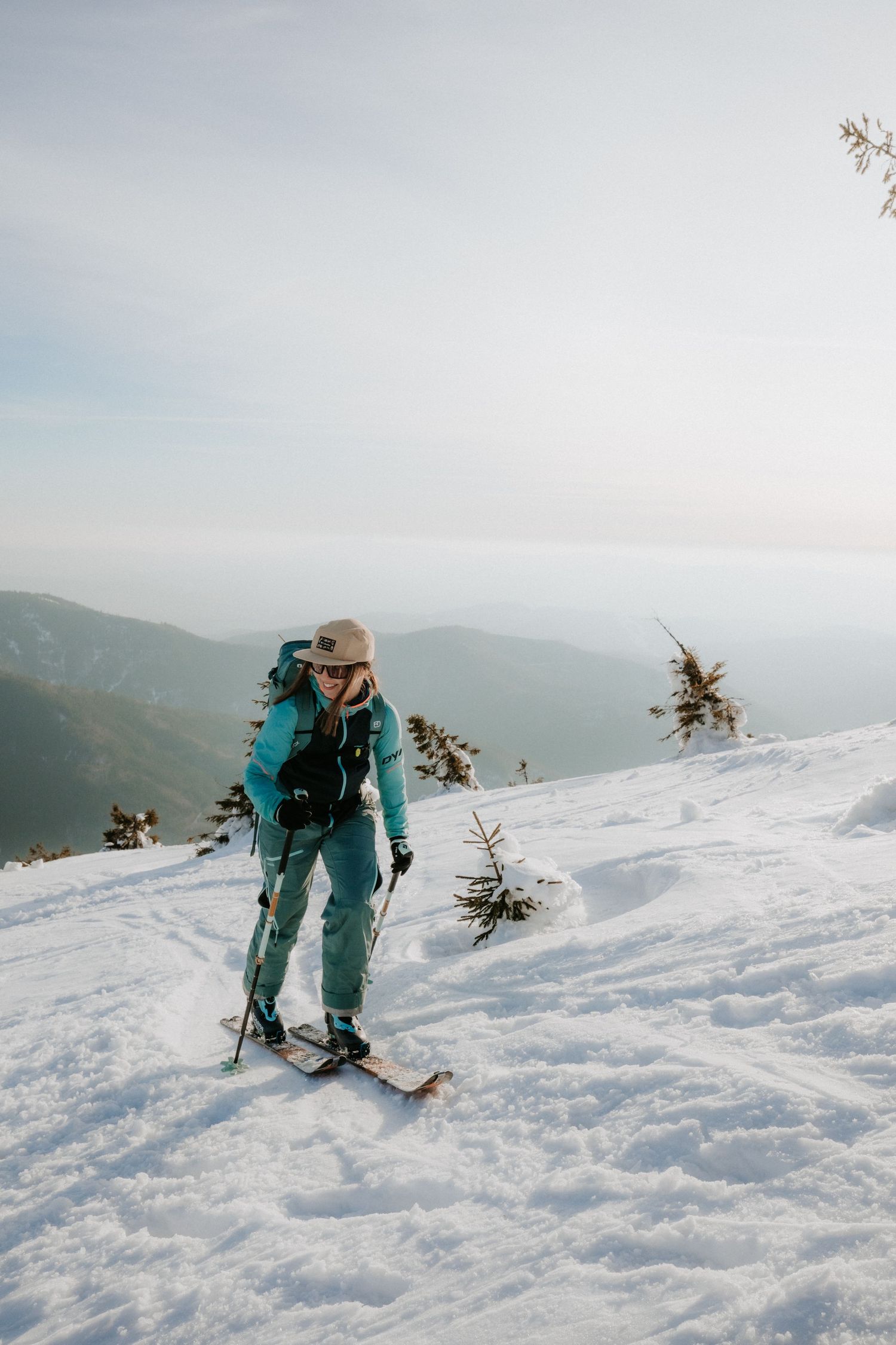 Female backcountry skier in teal jacket and tan cap ascending a snowy mountain slope with ski poles.