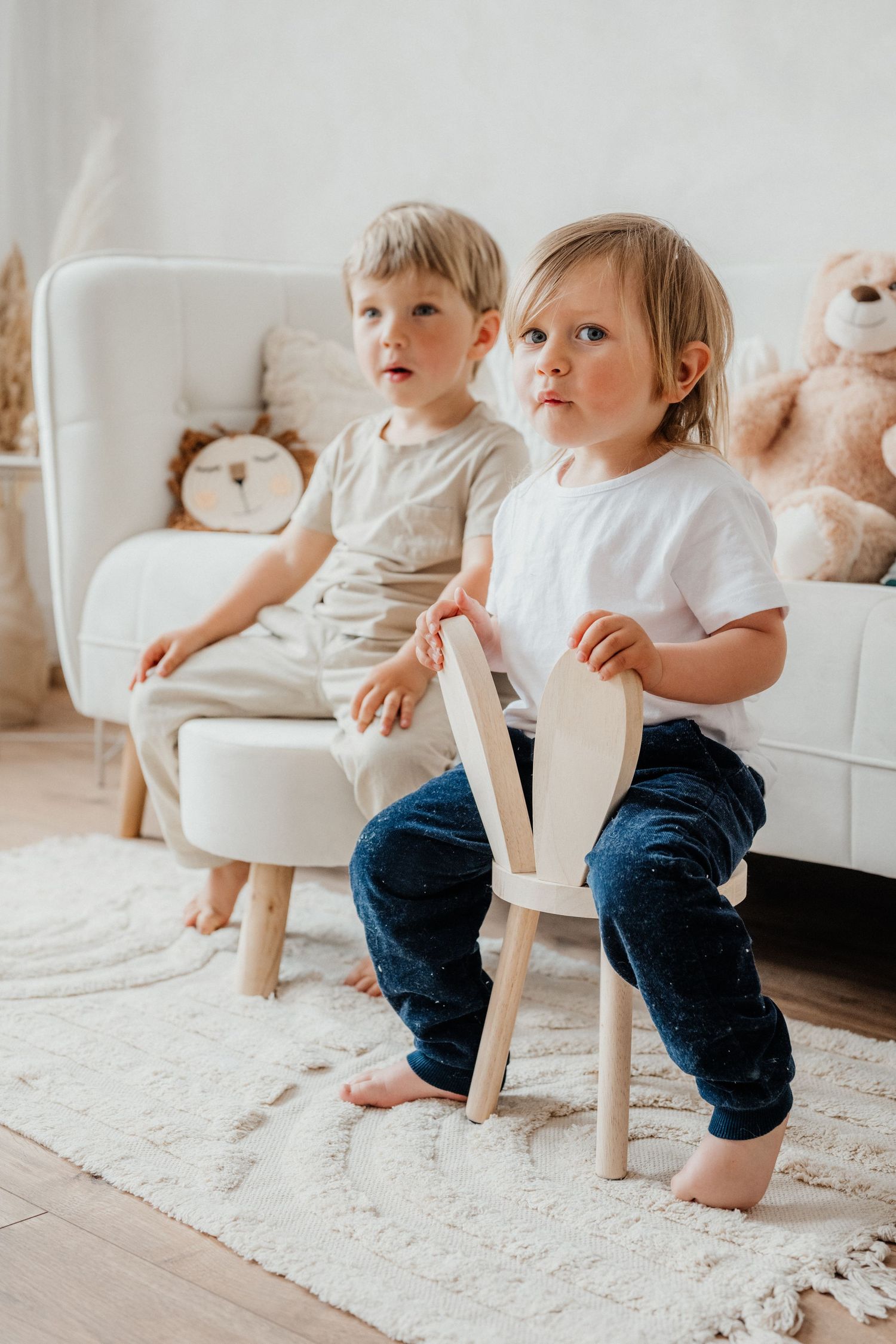 Two barefoot toddlers sitting on chairs in a bright living room with a white sofa.