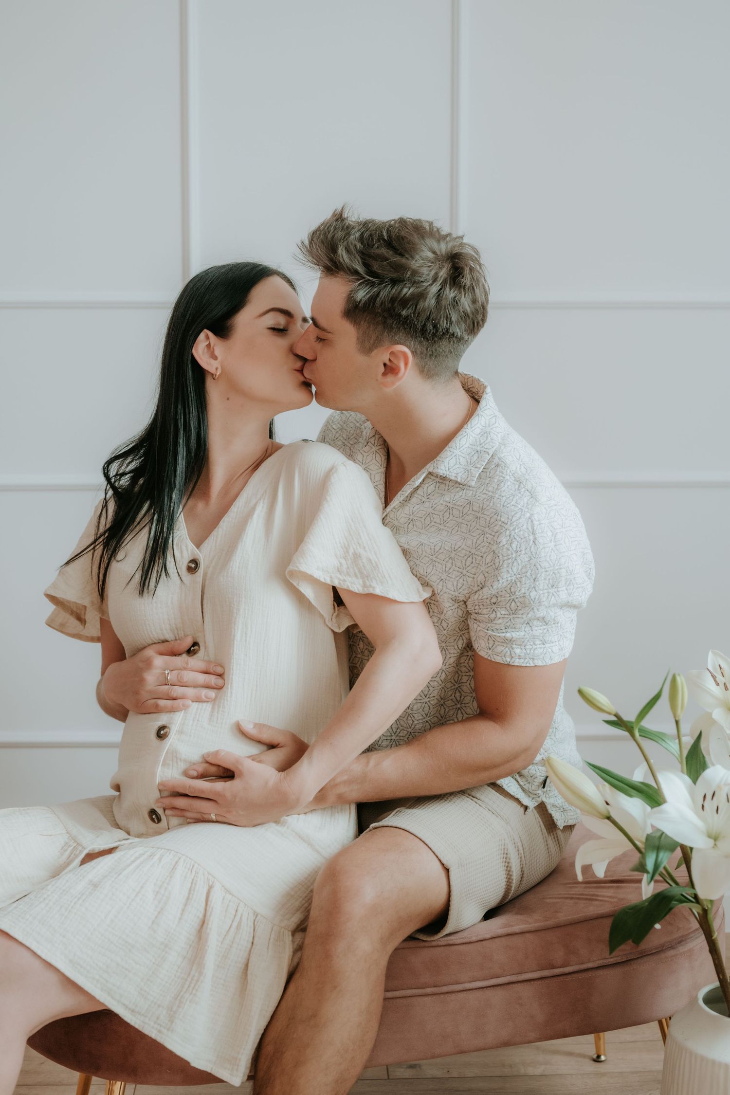 Couple kissing on pink velvet bench, man holding pregnant womans belly, white lilies nearby.