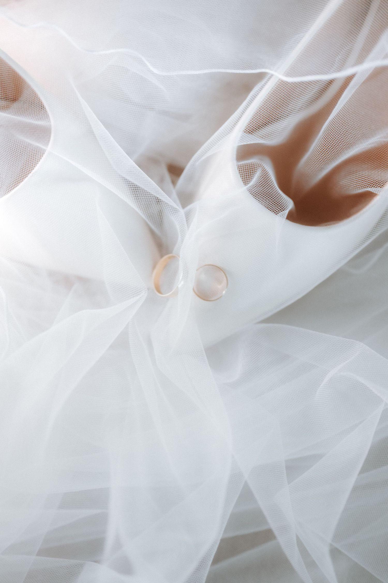 Two gold wedding rings resting on white bridal shoes and soft tulle veil fabric.
