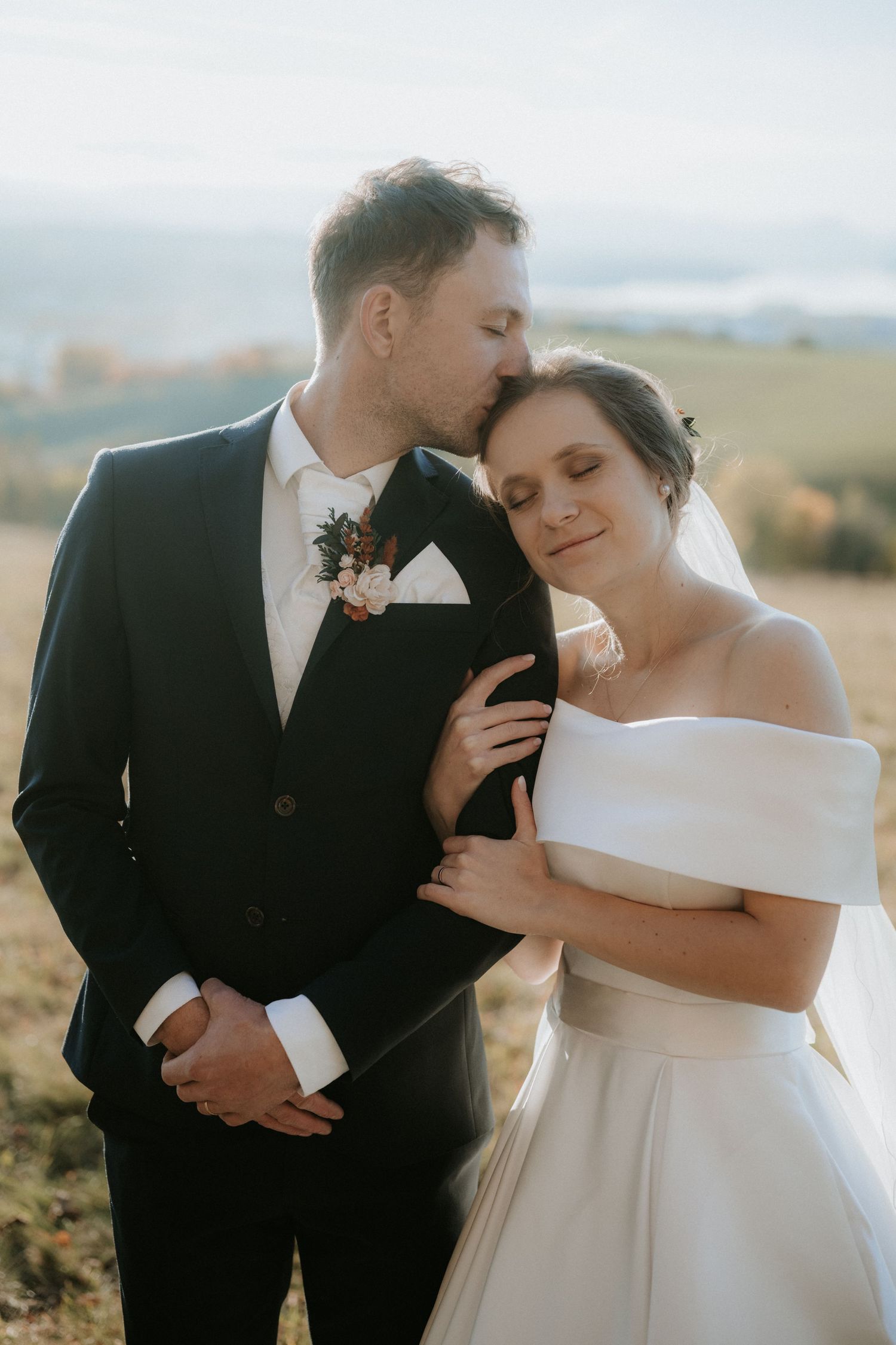 Groom in black suit kissing bride in off-shoulder white gown on a sunlit mountain meadow.