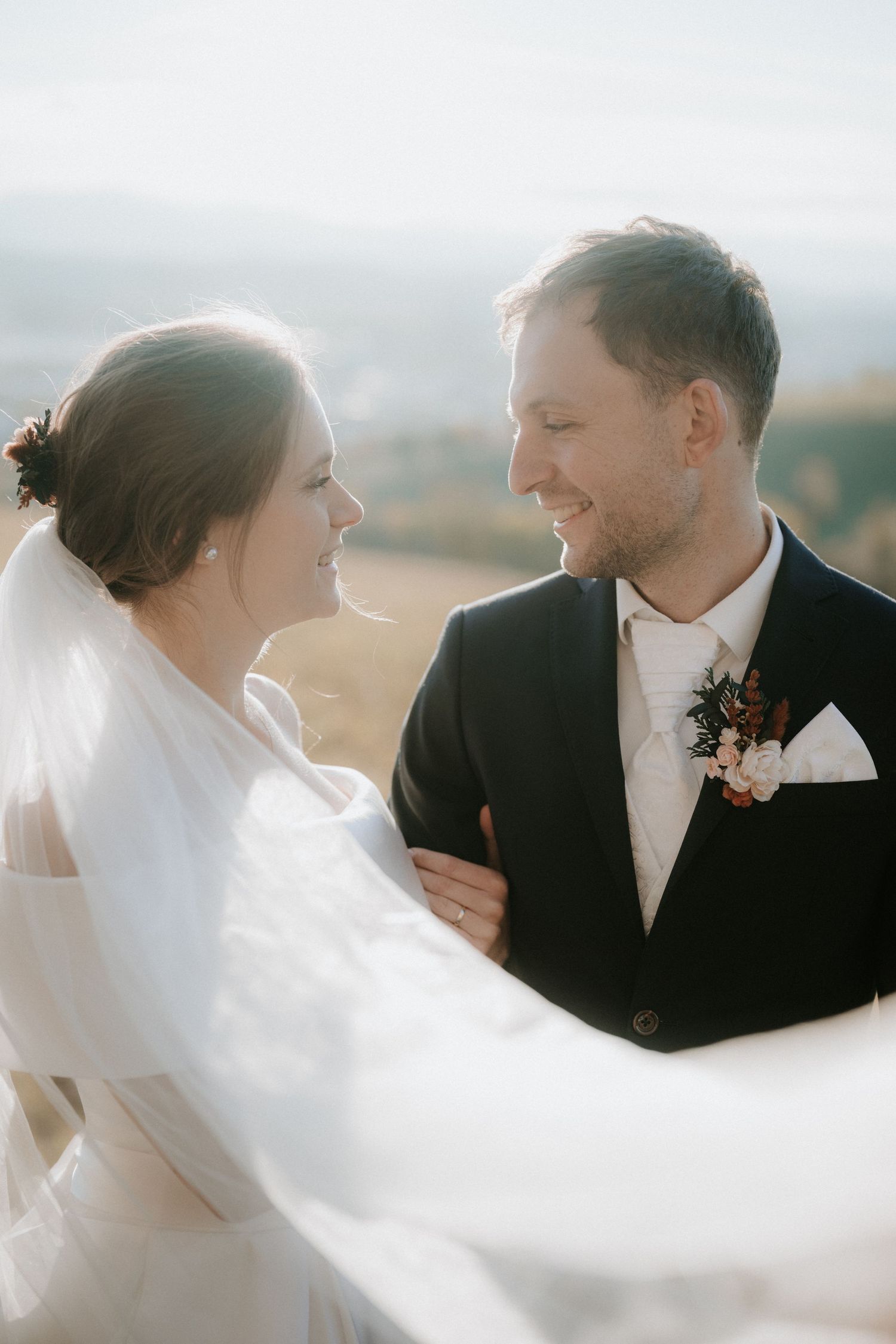 Bride and groom smiling at each other outdoors with veil flowing in the wind on their wedding day.