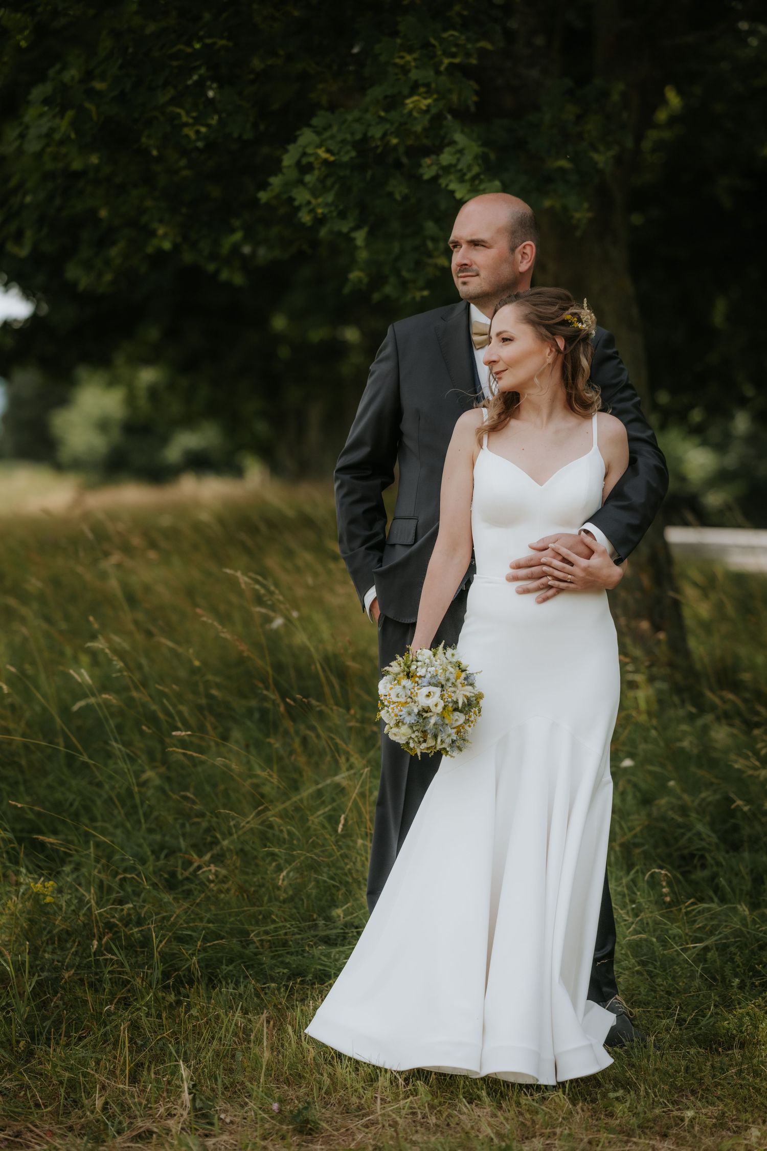 Bride in white gown holding bouquet embraced by groom in dark suit, standing in lush green field near trees.