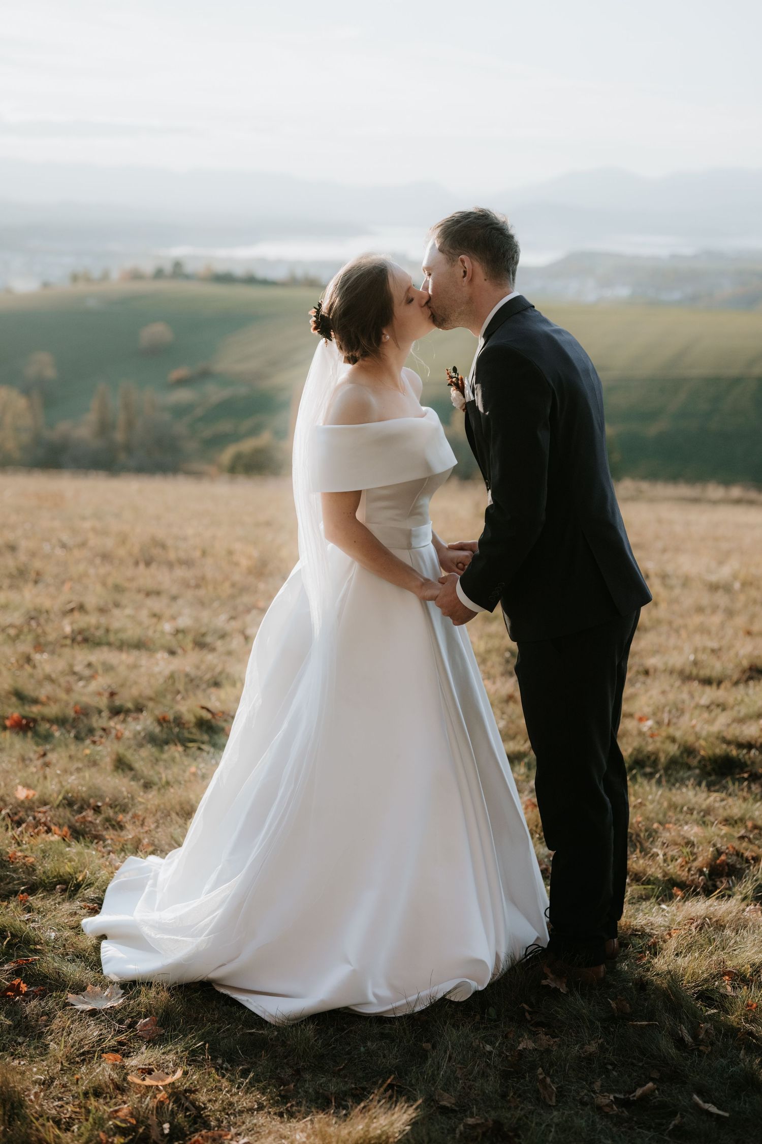 A bride and groom share a romantic kiss in a scenic countryside field, with rolling green hills and misty mountains in the background.