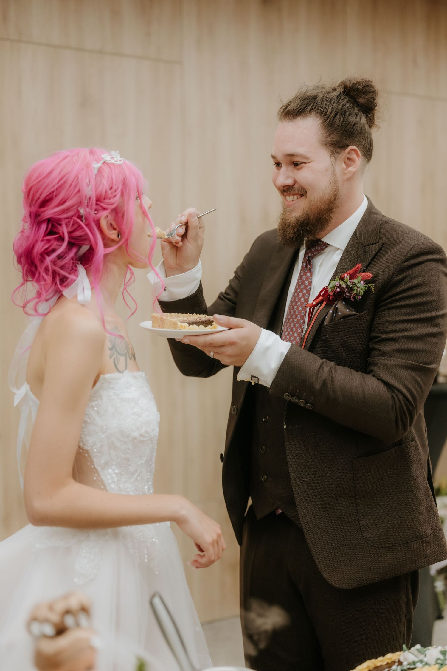 Groom feeding wedding cake to pink-haired bride at reception, both smiling joyfully in formal attire.