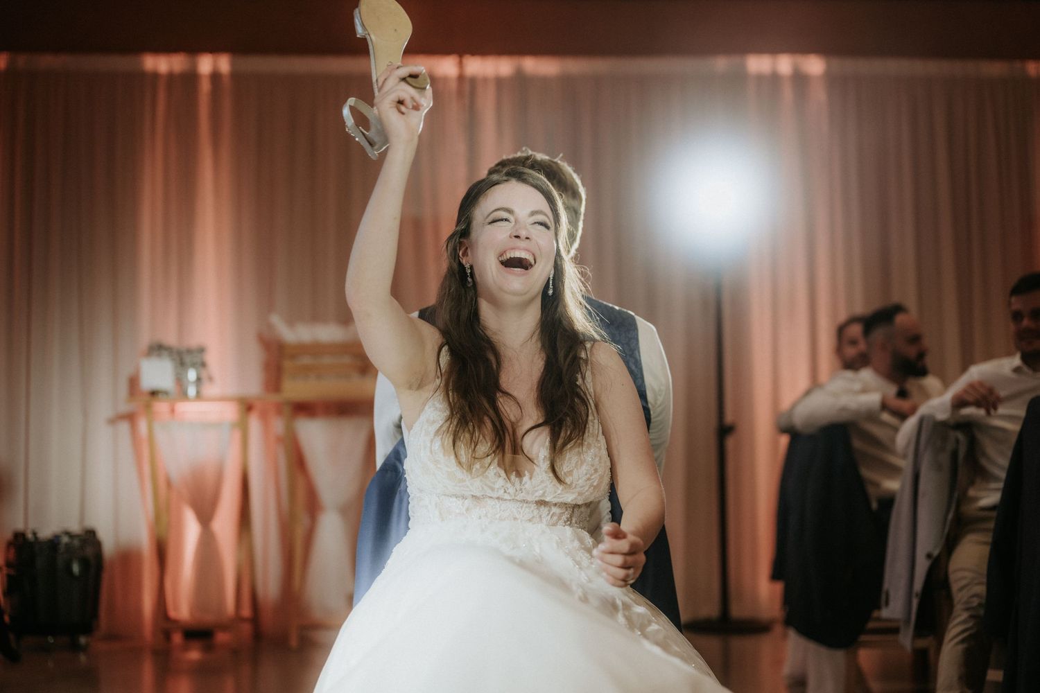 Bride in white gown laughing joyfully while raising her hand at a wedding reception celebration.