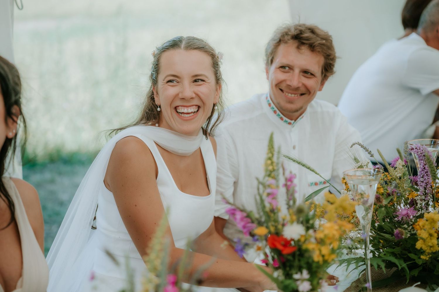 Smiling bride in white dress laughing at outdoor wedding reception with colorful wildflower arrangements.