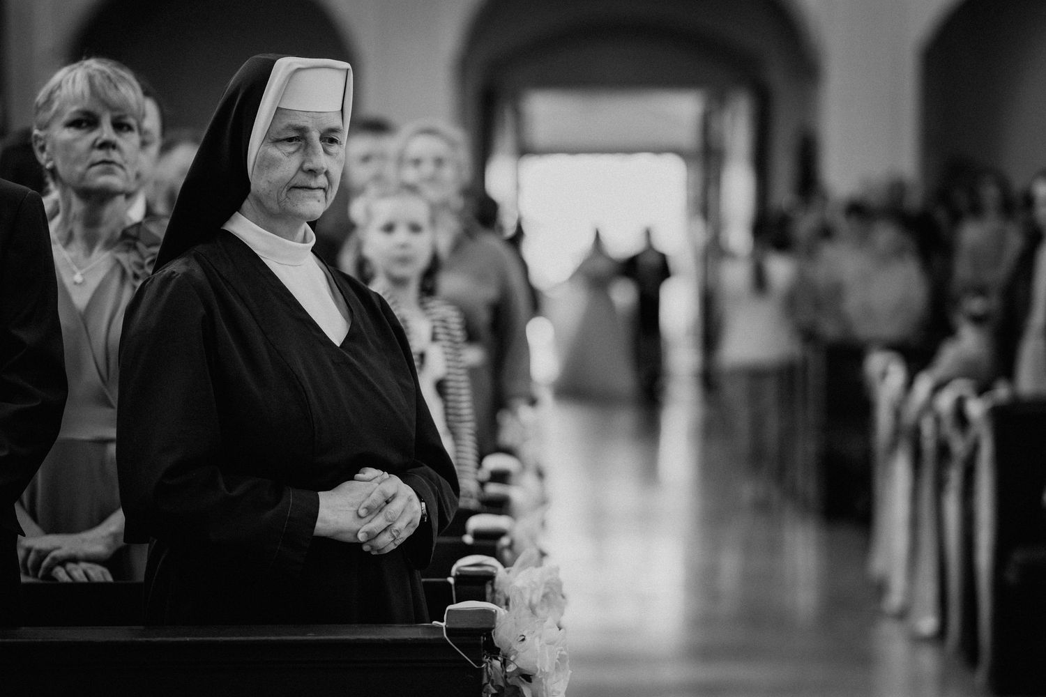 A nun in traditional black and white habit stands in a church aisle during a religious ceremony.