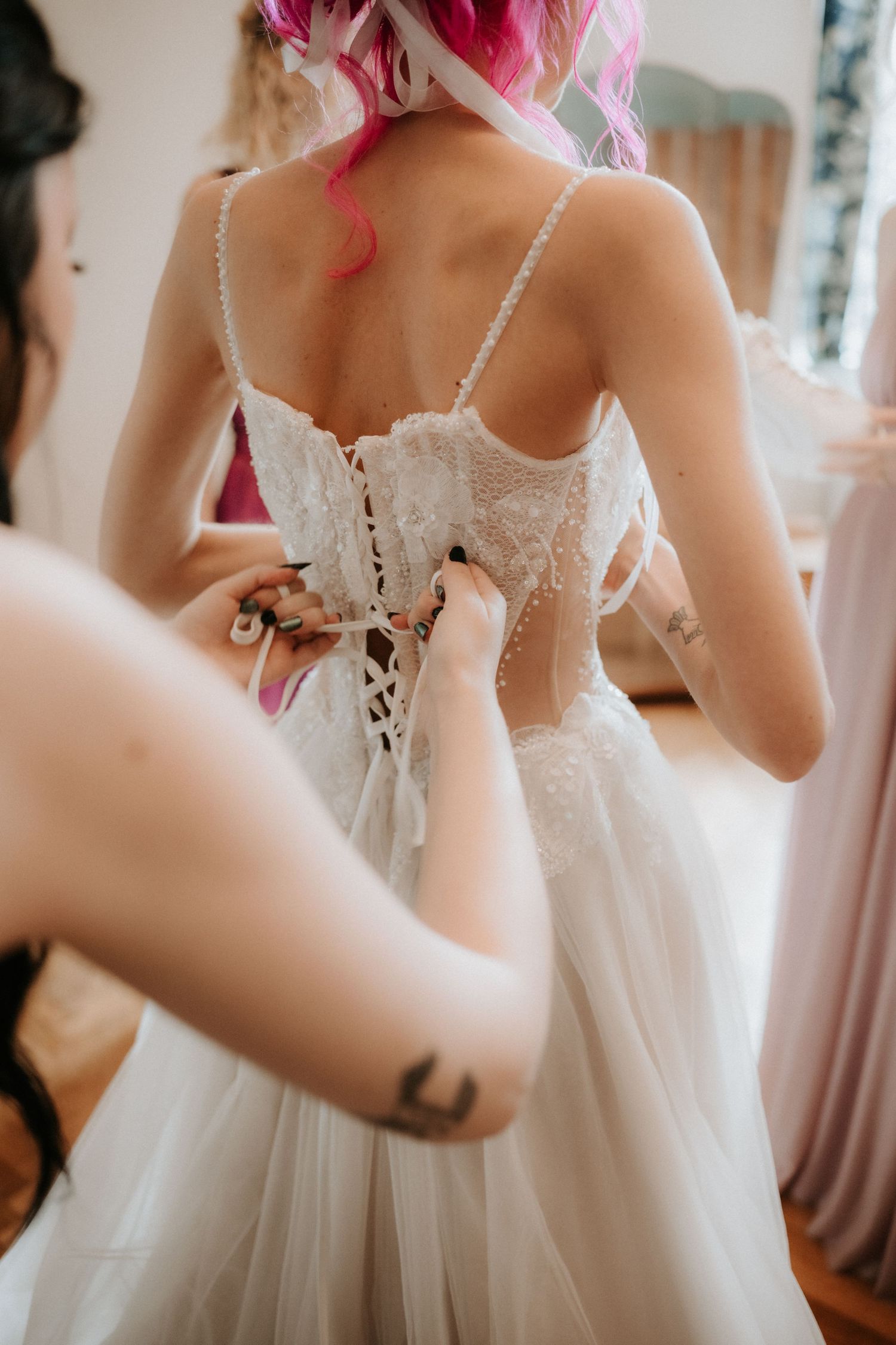 Bridesmaid lacing up the back of a white lace corset wedding dress on a bride with pink hair.