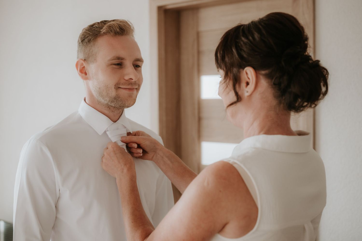A woman adjusts a mans white dress shirt collar as he smiles softly in a bright room.