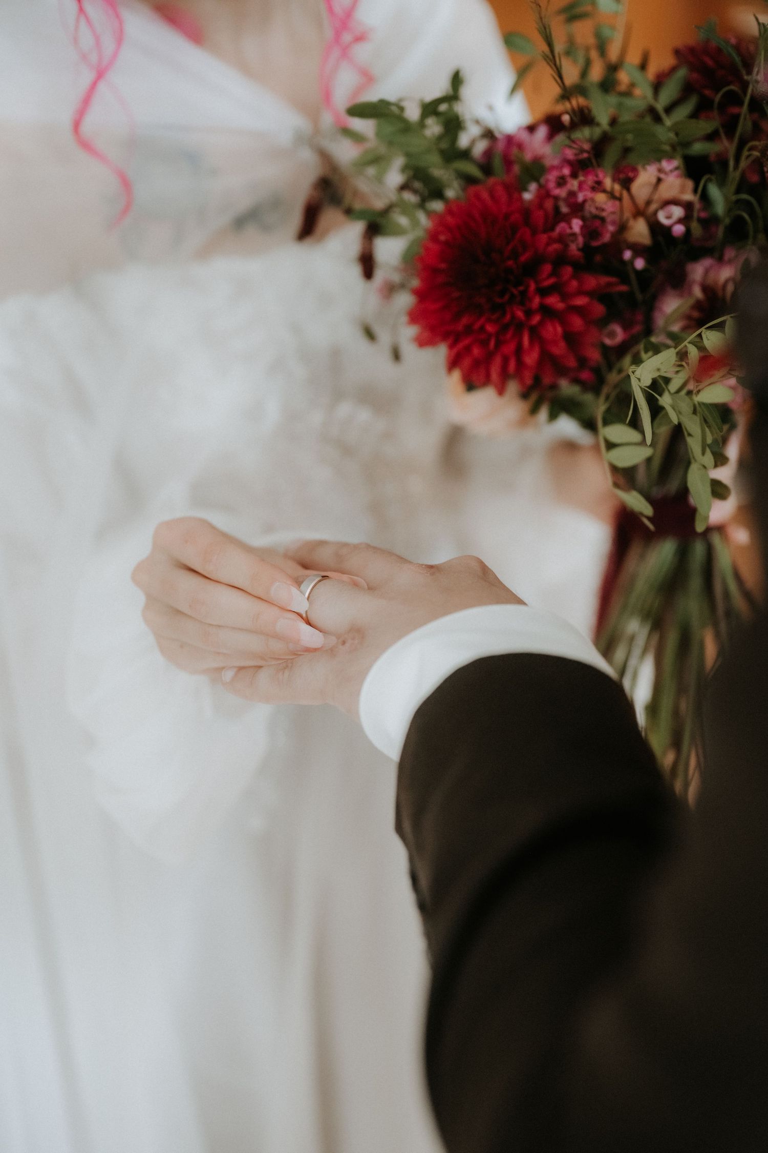 Groom holding bride's hand with wedding ring, beside a red floral bouquet with dahlias and greenery.