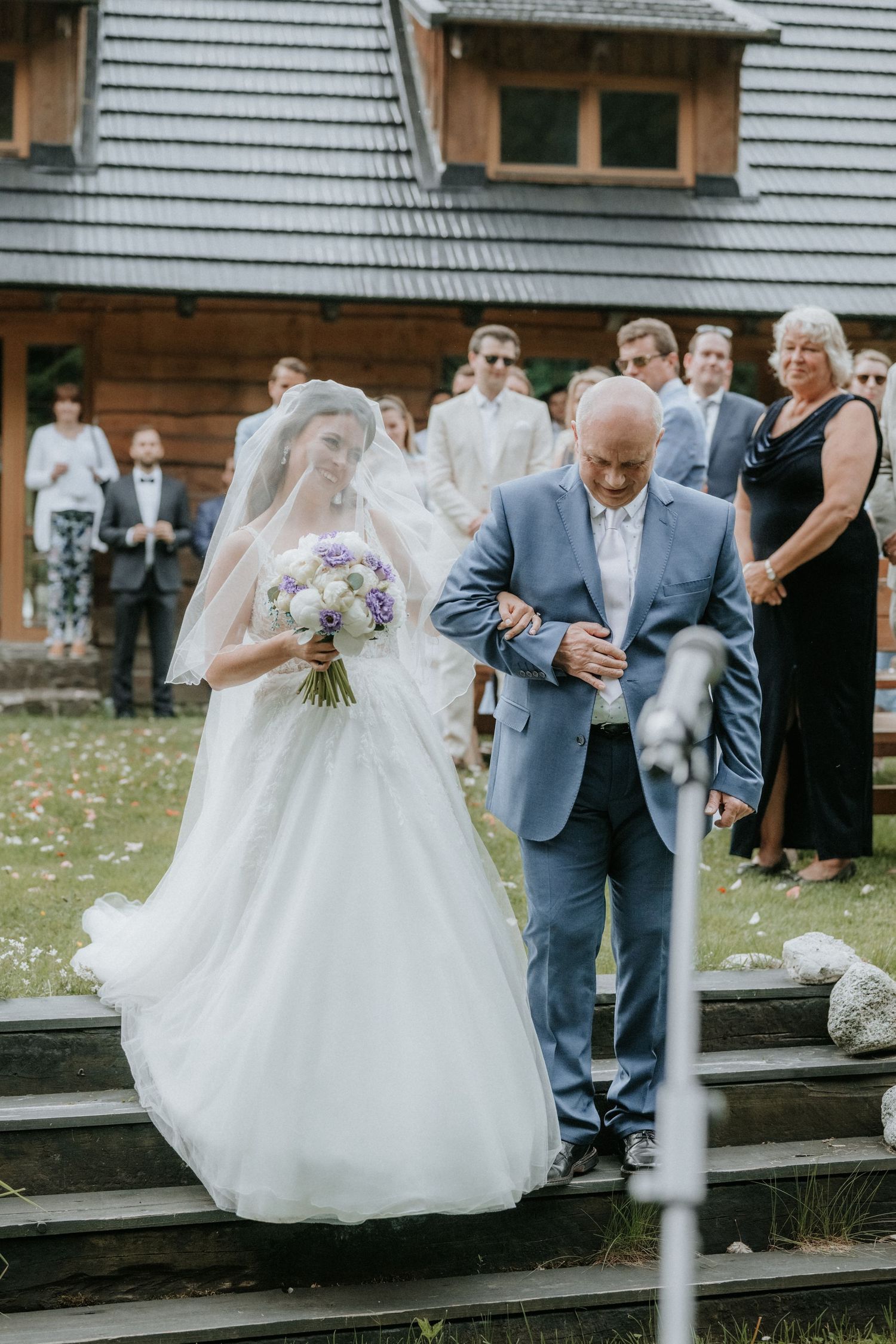 Bride in white gown with veil holding purple bouquet walks with man in blue suit down steps at outdoor wedding ceremony.