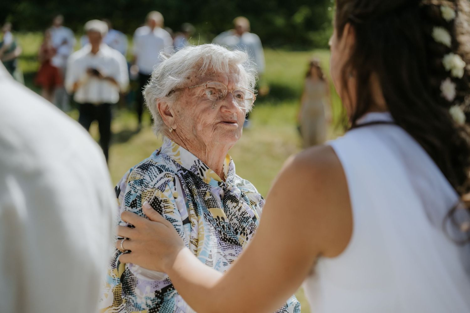 Elderly woman dancing outdoors at a garden celebration, smiling warmly while holding hands with a younger woman in white.