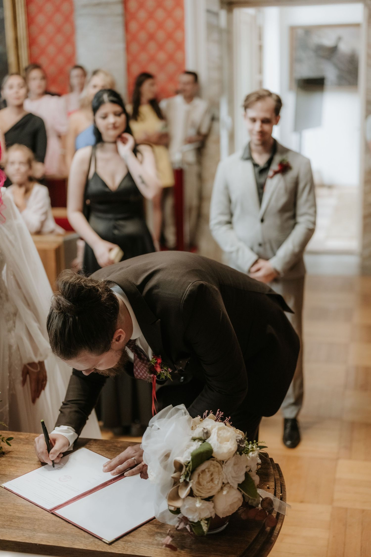 Groom signing wedding registry document at ceremony while guests watch in elegant venue with bridal bouquet nearby.