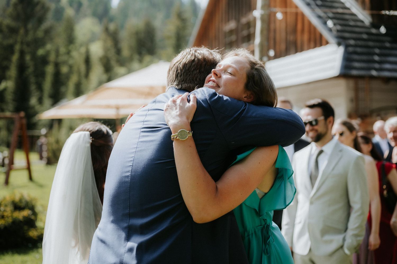 Emotional wedding hug between groom in navy suit and guest in teal dress outdoors at mountain venue.