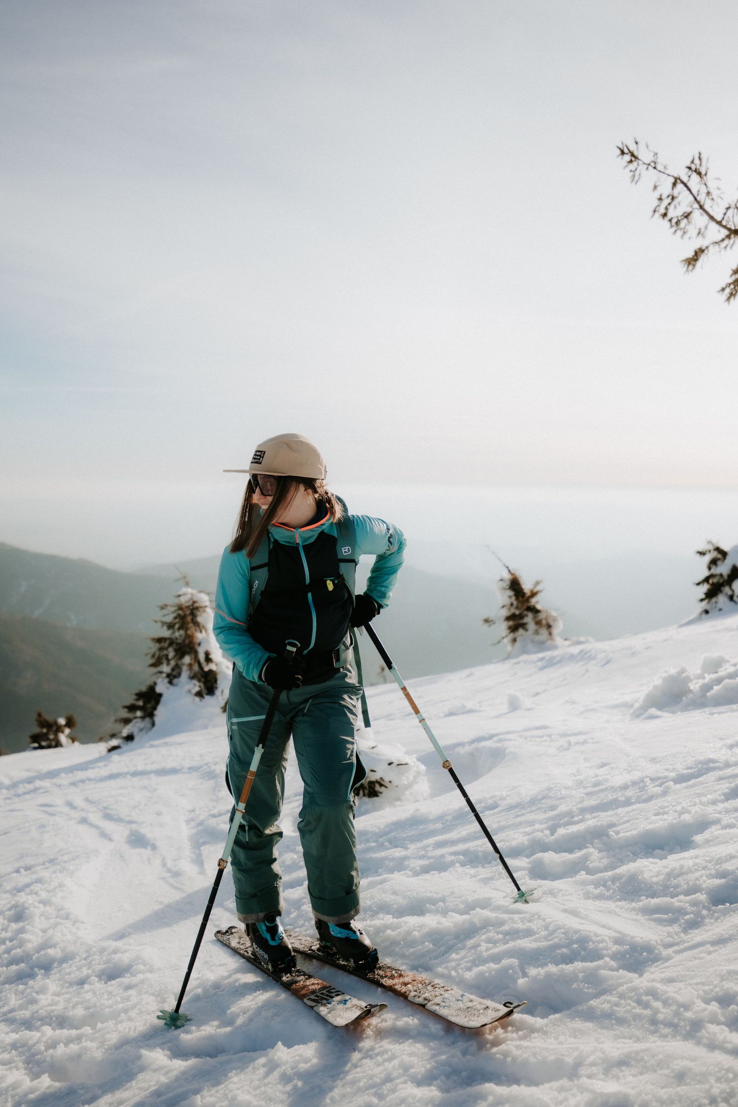Woman backcountry skiing on a snowy mountain ridge, adjusting her gear while holding ski poles.