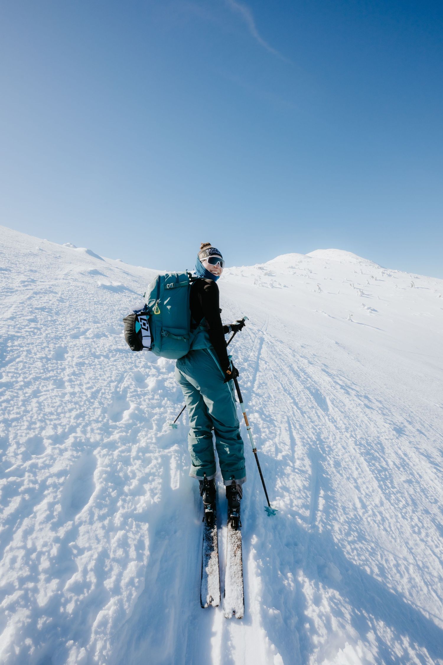 A skier with teal jacket and backpack climbs a snowy mountain slope on a clear sunny day.