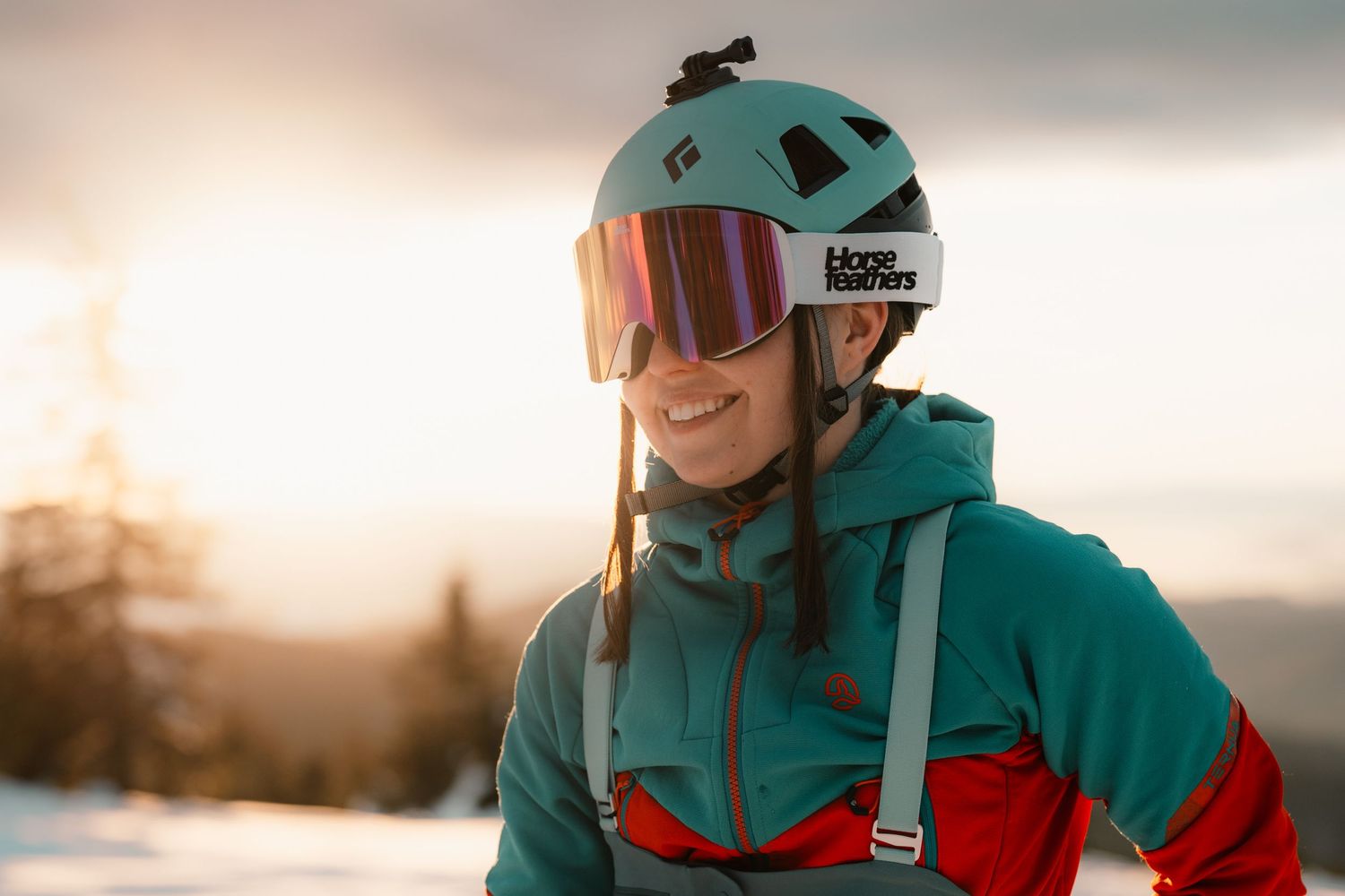 A smiling skier wearing a teal helmet and goggles with a Blore Fischer logo at a snowy sunset resort.