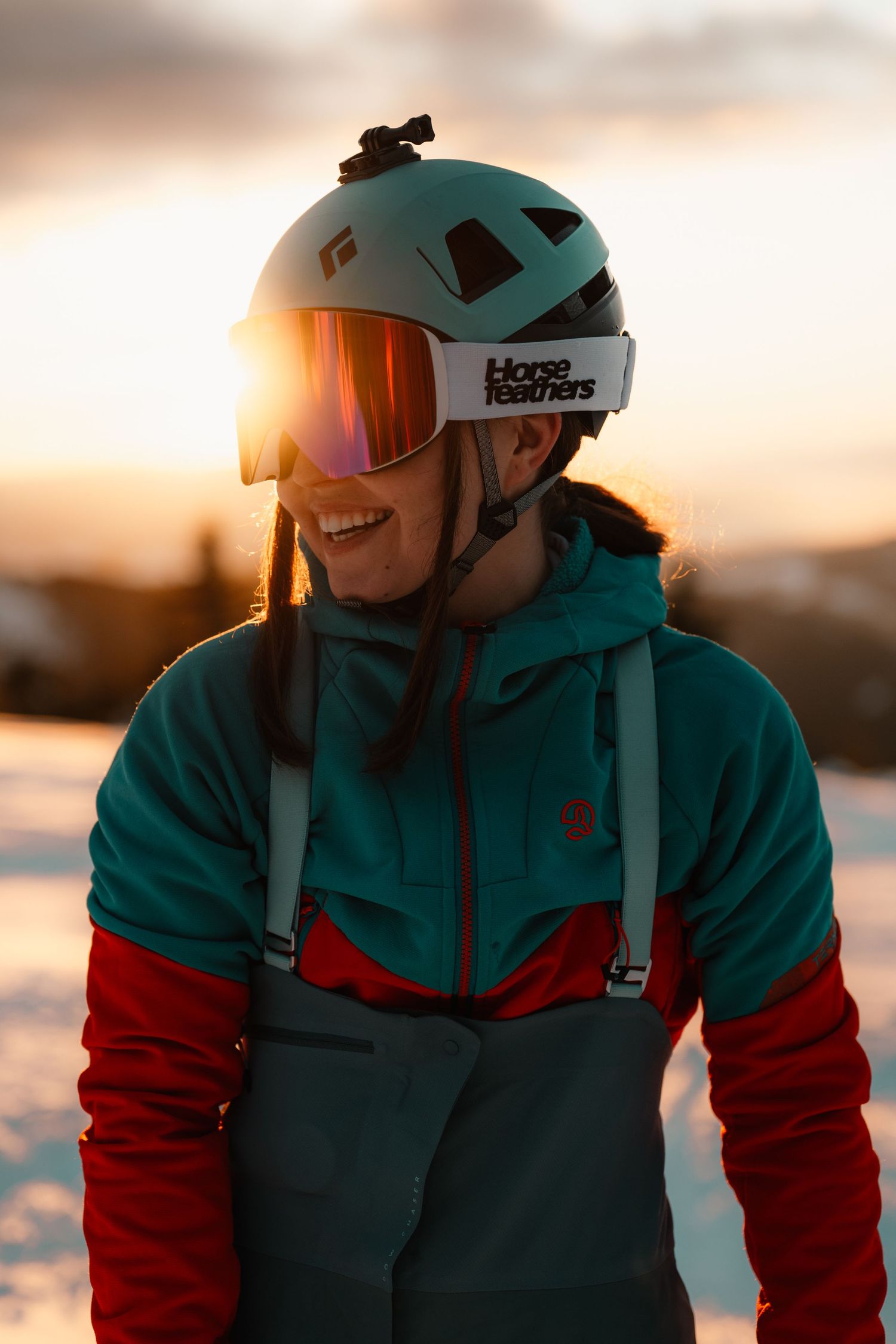 Happy female skier wearing Horse Feathers goggles and teal helmet glowing in golden sunset light.