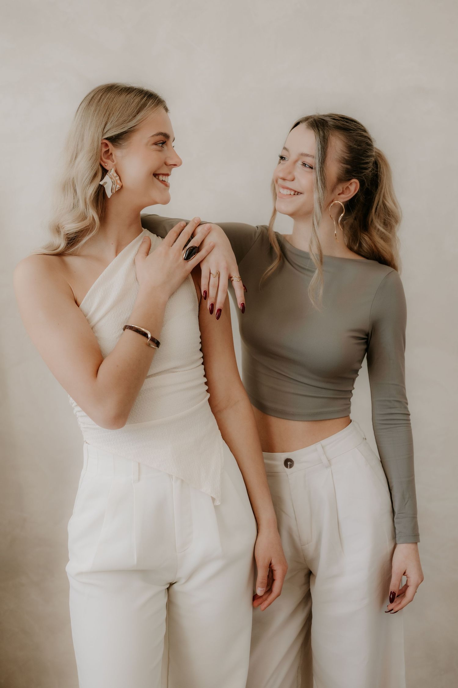 Two smiling women in neutral outfits laughing together against a beige textured wall.