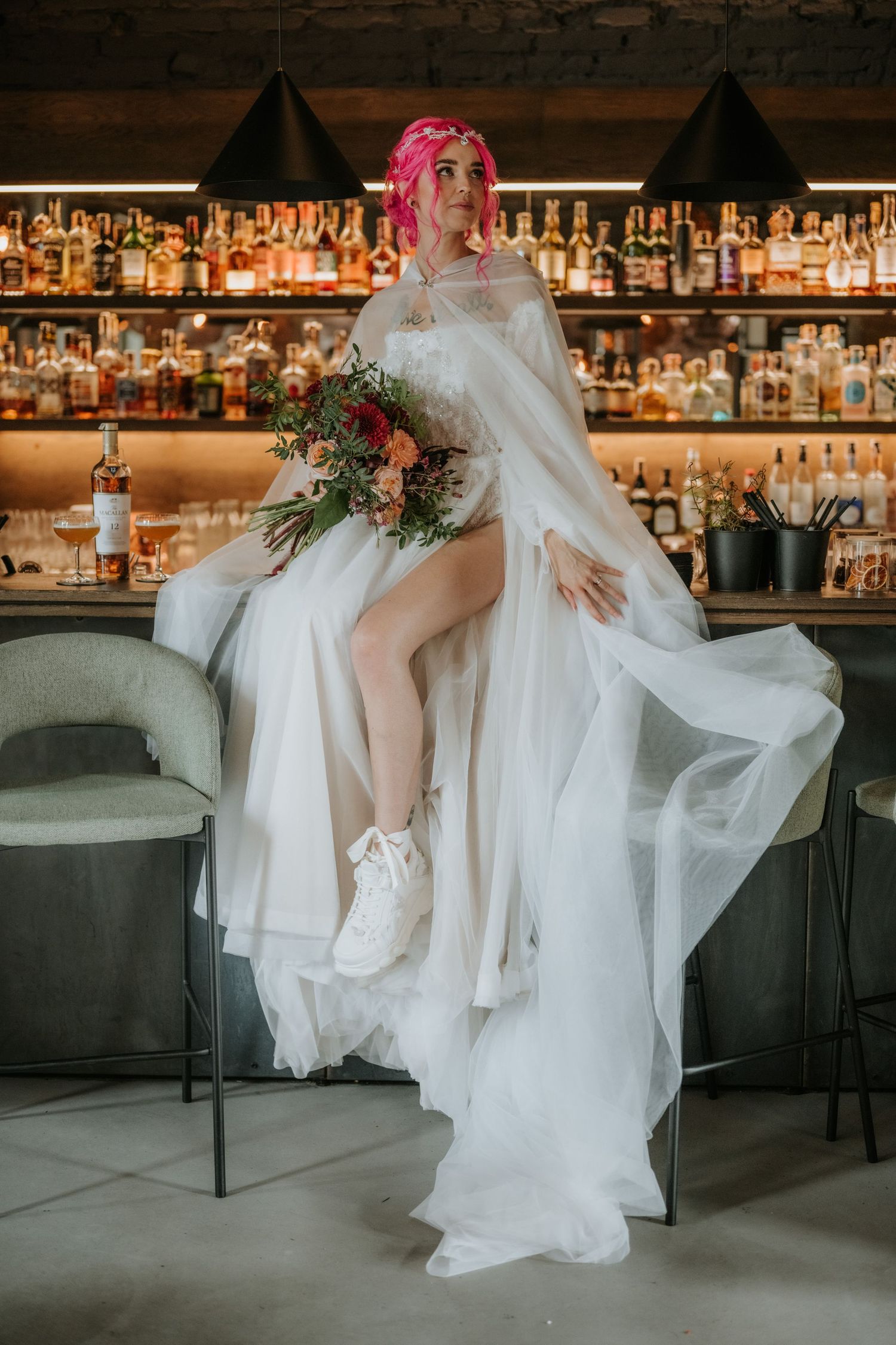 Pink-haired bride in white gown and tulle cape sitting at bar holding colorful bridal bouquet.