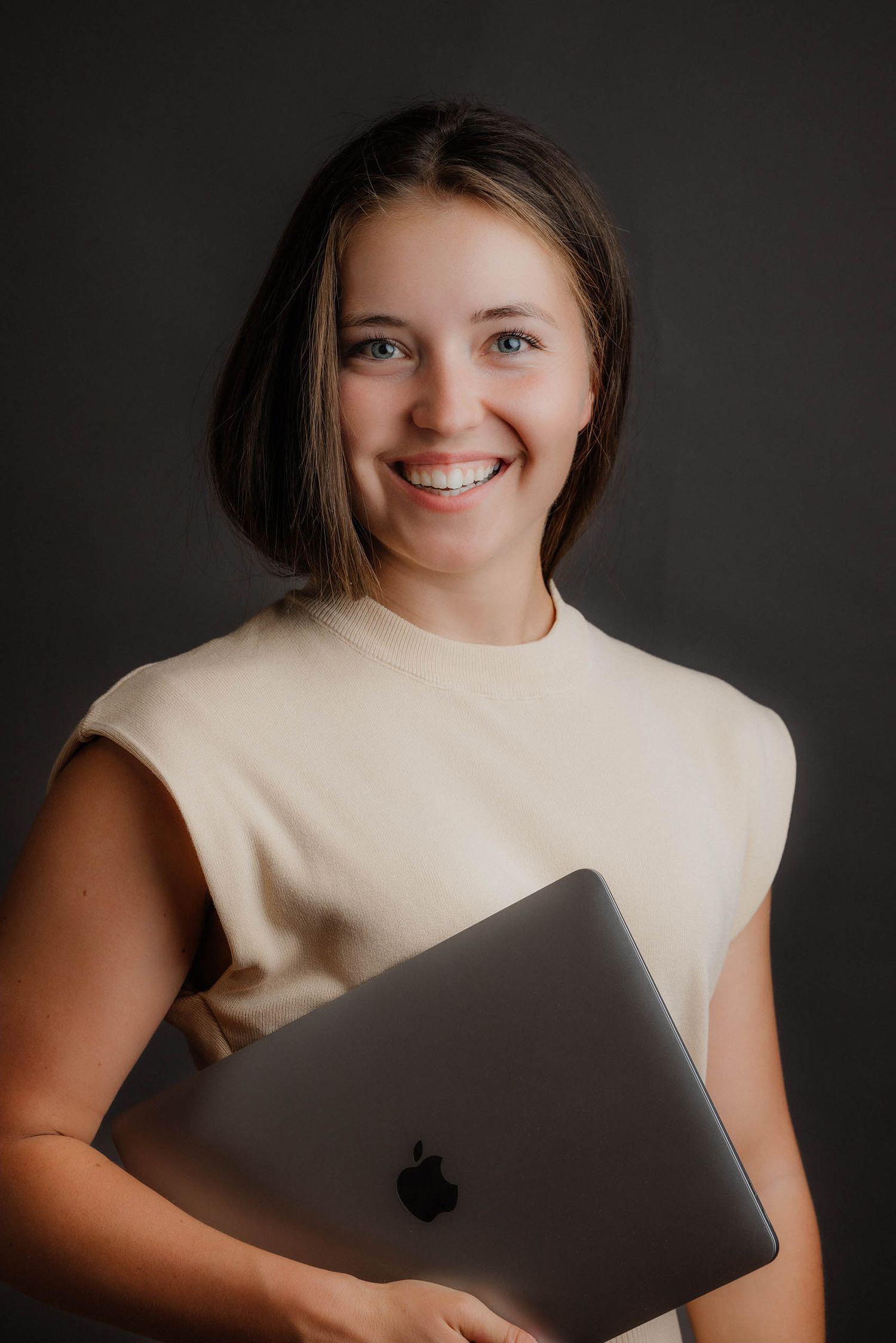 Smiling young woman holding Apple MacBook laptop against dark gray background.